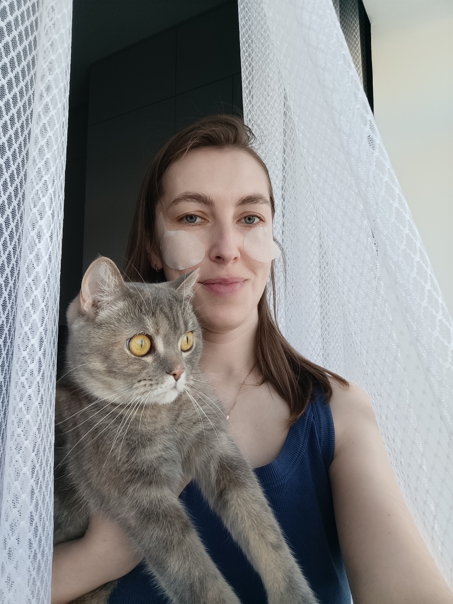 A woman with fair skin and brown hair is holding a gray tabby cat with yellow eyes. She is wearing light-colored eye patches under each eye and a blue sleeveless top. They are positioned in front of a window with a white lace curtain. To the left of the window is a dark gray wall or cabinet. The woman is smiling slightly and looking towards the camera. The cat is looking to the left of the frame.
This image was taken in Chaikovsky, Russia.