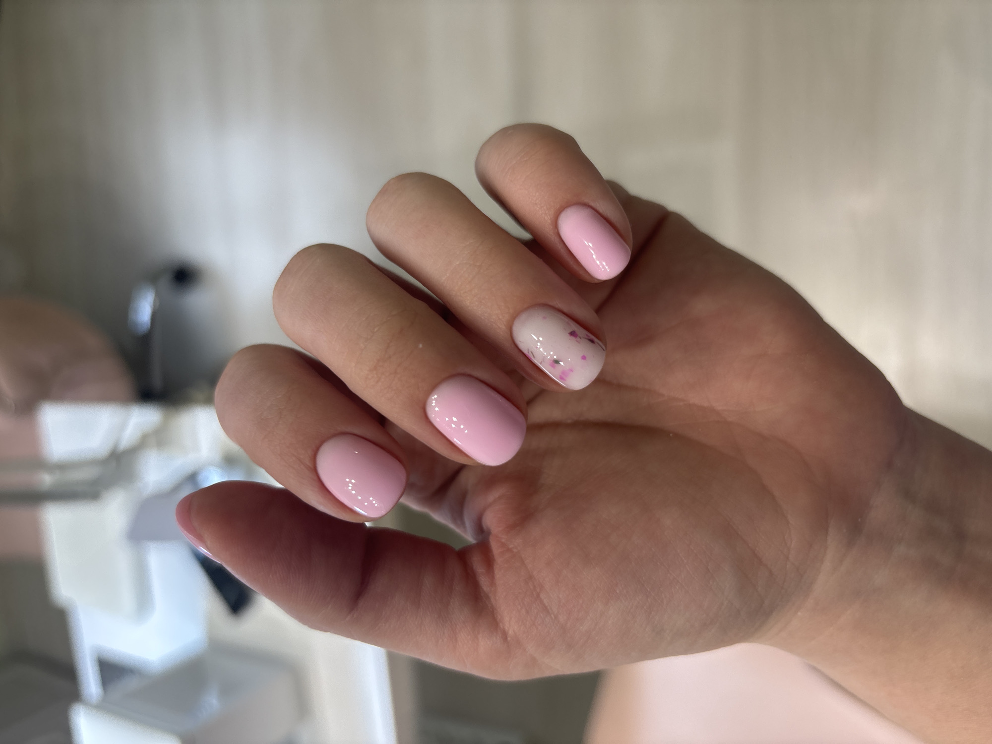 A close-up vertical shot displays a left human hand, located in the city of Ферма, Russia. The hand is positioned with fingers slightly spread. The fingernails are short to medium length and oval-shaped, exhibiting a fresh manicure. The thumb, index, middle, and pinky fingers are coated with a solid, glossy, light pink nail polish. The ring finger features a milky white or translucent base coat with small, scattered pink-reddish glitter particles embedded within. The skin of the hand appears clear and smooth. The background is significantly out of focus, presenting a light-colored, indistinct surface, possibly a tabletop, and blurred elements consistent with indoor furniture or equipment, such as a white rectangular object on the left.
