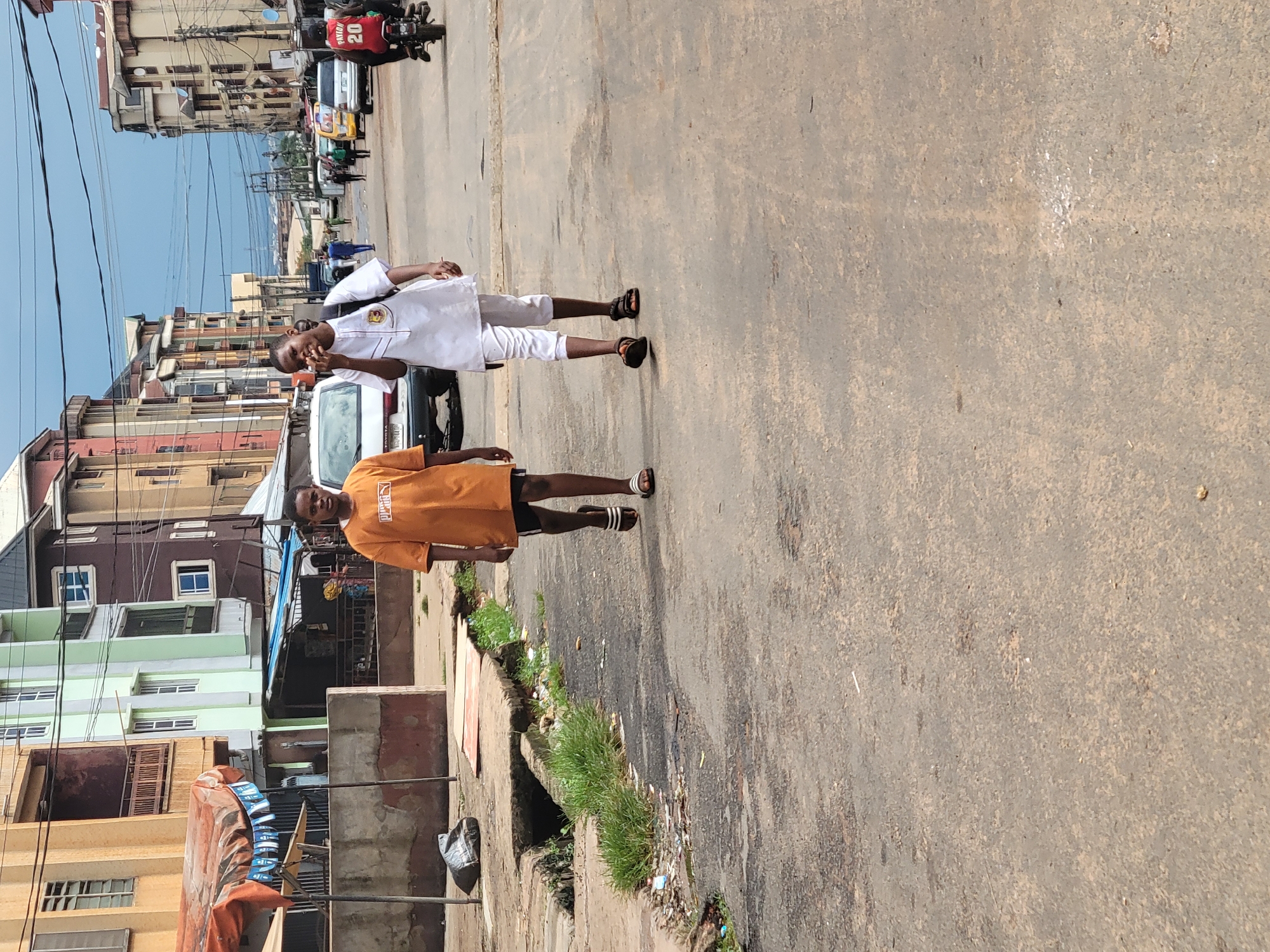 This is a medium-shot, eye-level, natural-light photograph taken on a street in Onitsha, Nigeria. Two young males are walking towards the camera from the middle ground of the scene. The male on the left is wearing an orange t-shirt and dark shorts, and appears to be looking at the camera. The male on the right is wearing a white traditional outfit and a backpack, and is looking towards the left and appears to be speaking or gesturing. They are walking on a paved street with a gutter to their left, which contains some grass and debris. In the background, there are multi-story buildings with balconies, and several vehicles, including a white sedan and a motorcycle with a rider wearing a red jersey. The sky is overcast. The scene depicts a typical urban environment with commercial and residential buildings lining a street.