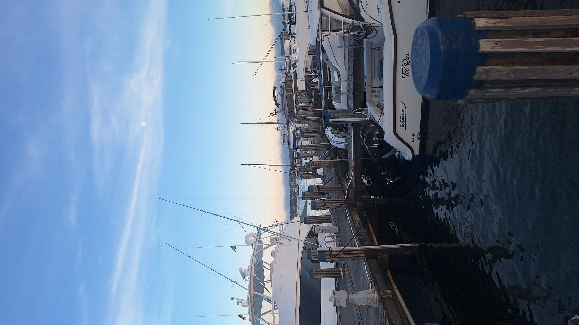 The image depicts a waterfront scene in Nantucket, Massachusetts, likely at a marina or harbor. Several boats are docked, ranging in size from smaller fishing vessels to larger yachts. The boats are moored to a wooden dock, and some are equipped with fishing rods. The water is calm, reflecting the blue sky and some scattered clouds. The sky has a soft, twilight glow, suggesting it might be early morning or late evening. The moon is visible high in the sky. In the foreground, a weathered wooden post topped with a blue cap stands next to the dock. The overall atmosphere is serene and nautical.