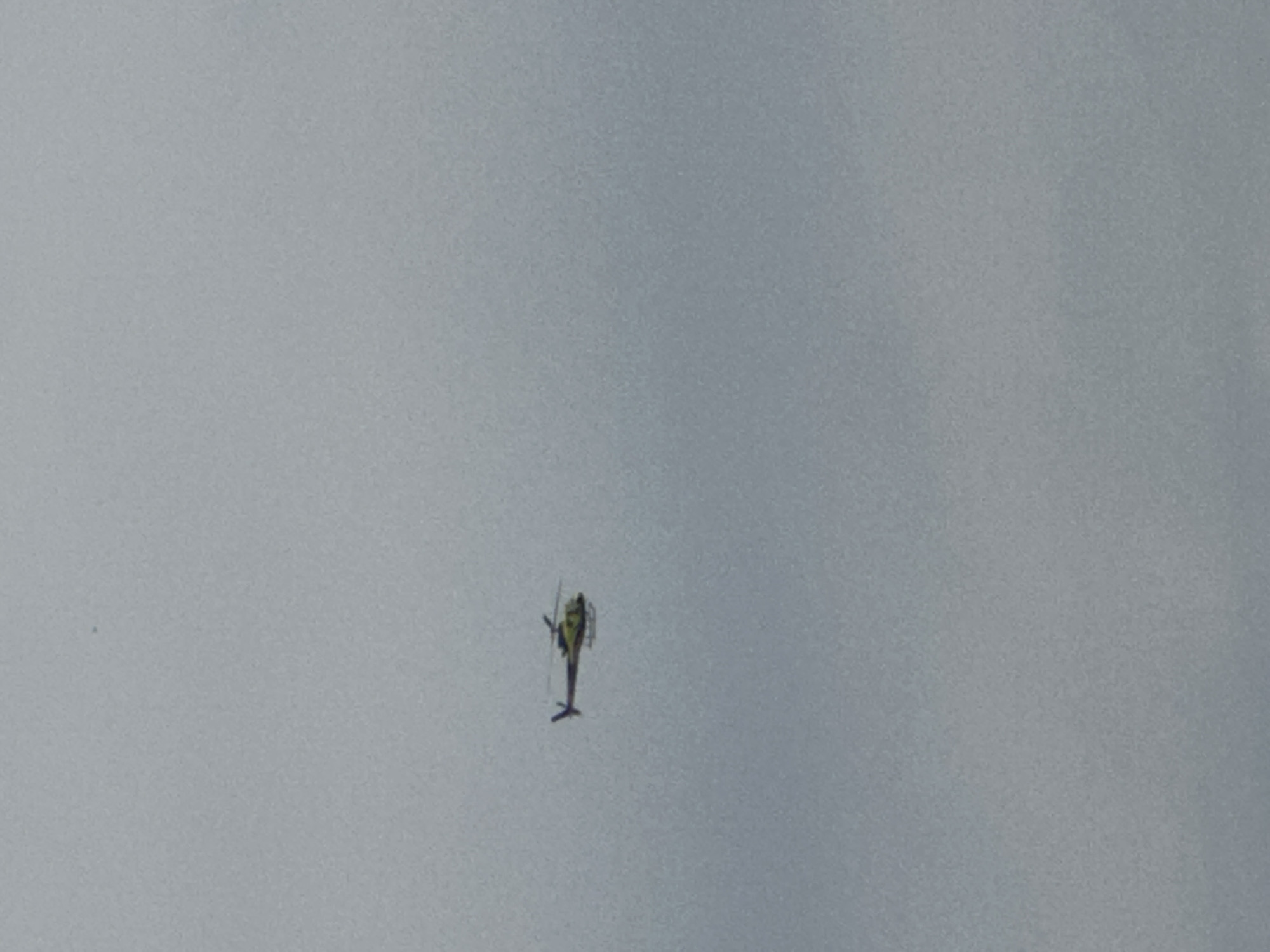 A single helicopter is captured in flight against an overcast sky in Korchivka, Ukraine. The helicopter is primarily yellow with dark rotor blades and tail rotor. It is angled with its nose pointing slightly upwards and to the left. The surrounding environment is uniformly gray and featureless, indicating a cloudy day. No people or other objects are visible in the frame.