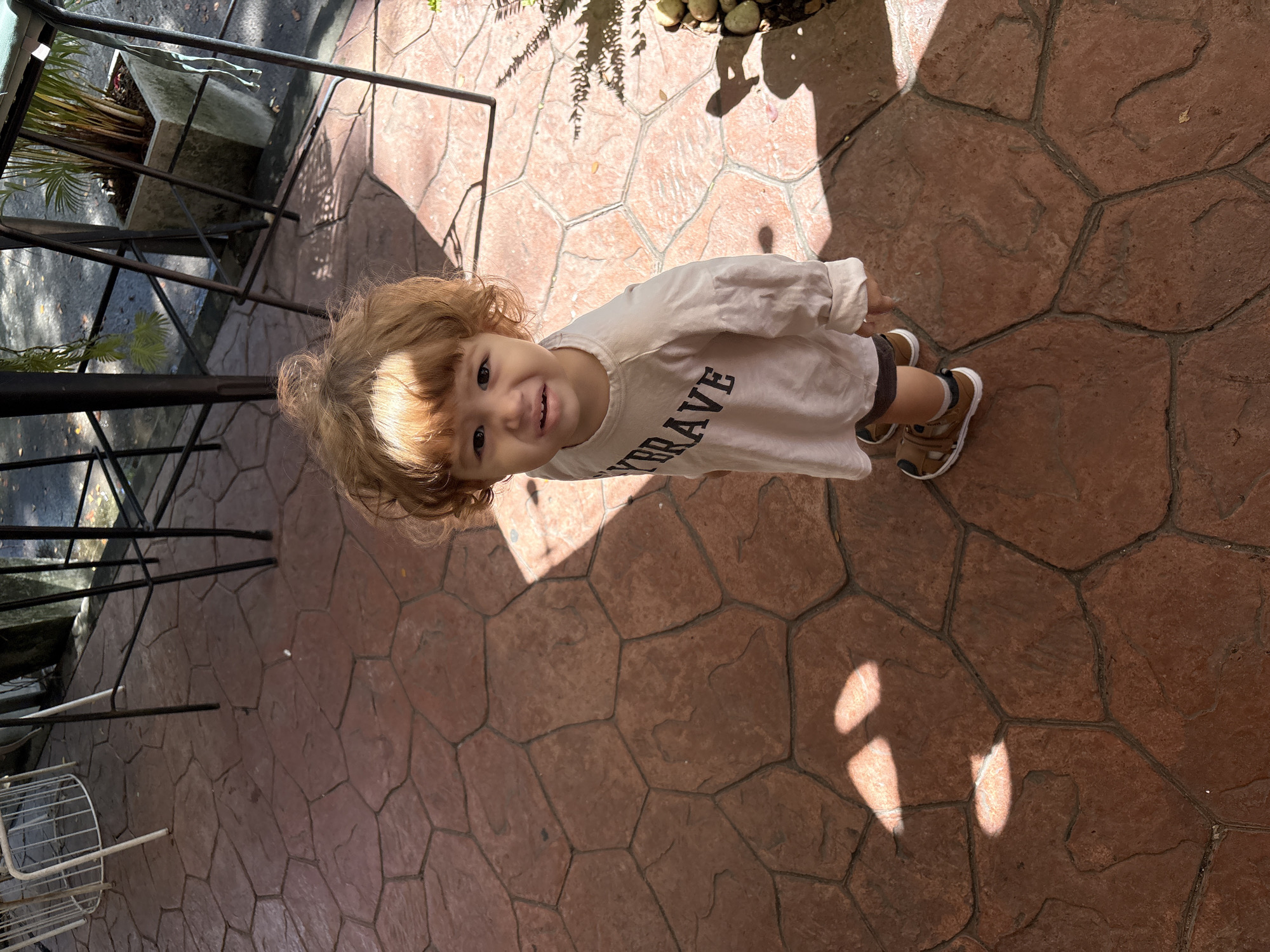 A young child with curly light brown hair stands on a red stamped concrete patio in Choeng Thale, Thailand. The child is wearing a long-sleeved white shirt with "BRAVE" printed in black lettering across the chest, and brown sandals with white soles. The child is looking up at the camera with a slightly distressed expression. The patio has a pattern of interlocking hexagonal and irregular shapes. Sunlight casts shadows from unseen objects onto the patio. In the background, metal chairs and a potted plant are visible.