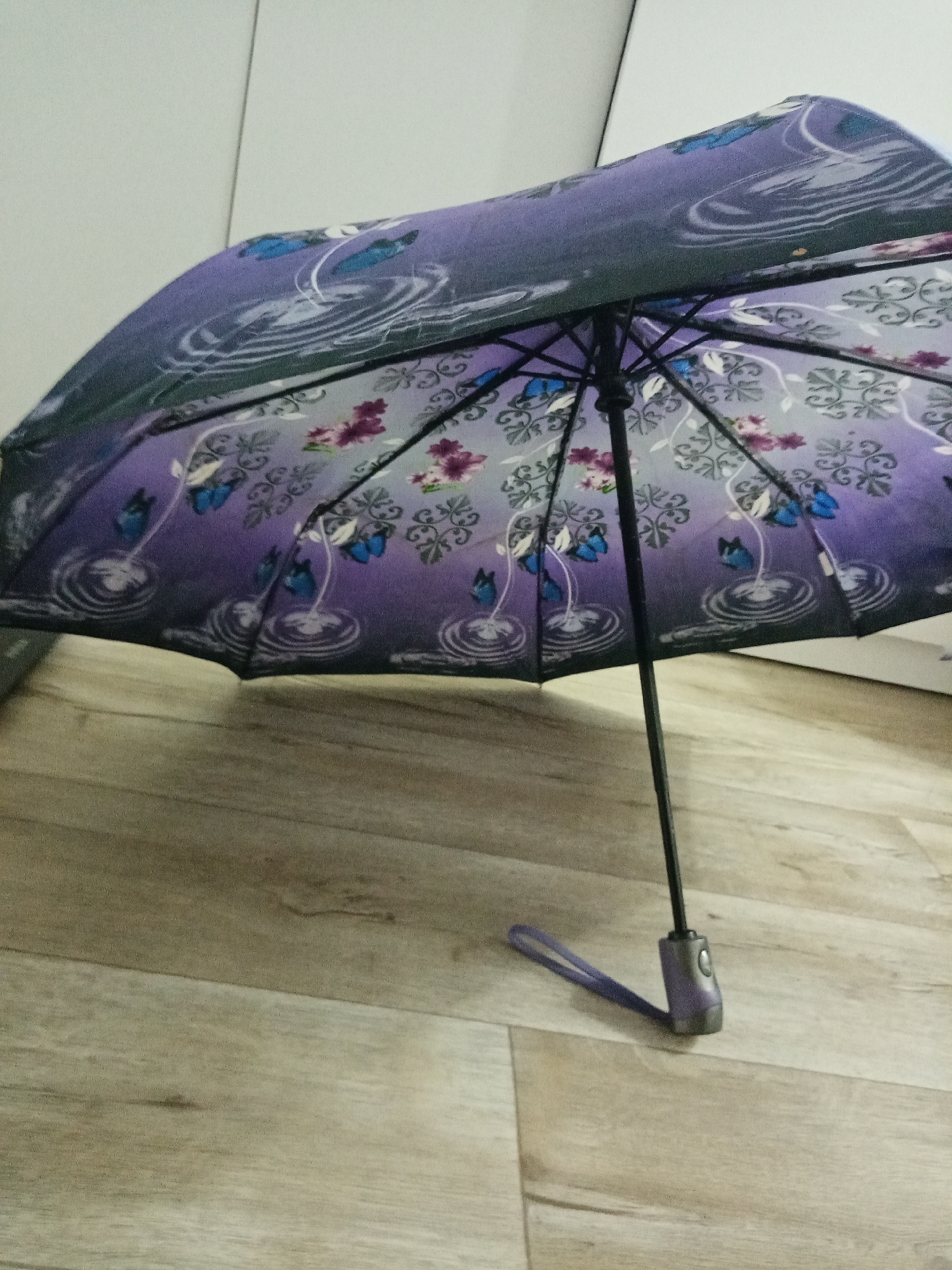 A slightly opened umbrella stands on a light-colored, wood-grain floor. The umbrella's fabric is a gradient of purple, adorned with butterfly and flower designs, and water ripple patterns. The umbrella frame is black with the handle and wrist strap visible. The background includes a white, possibly cabinet or wall, surface. The location in Elektrostal, Russia cannot be determined from the image alone.