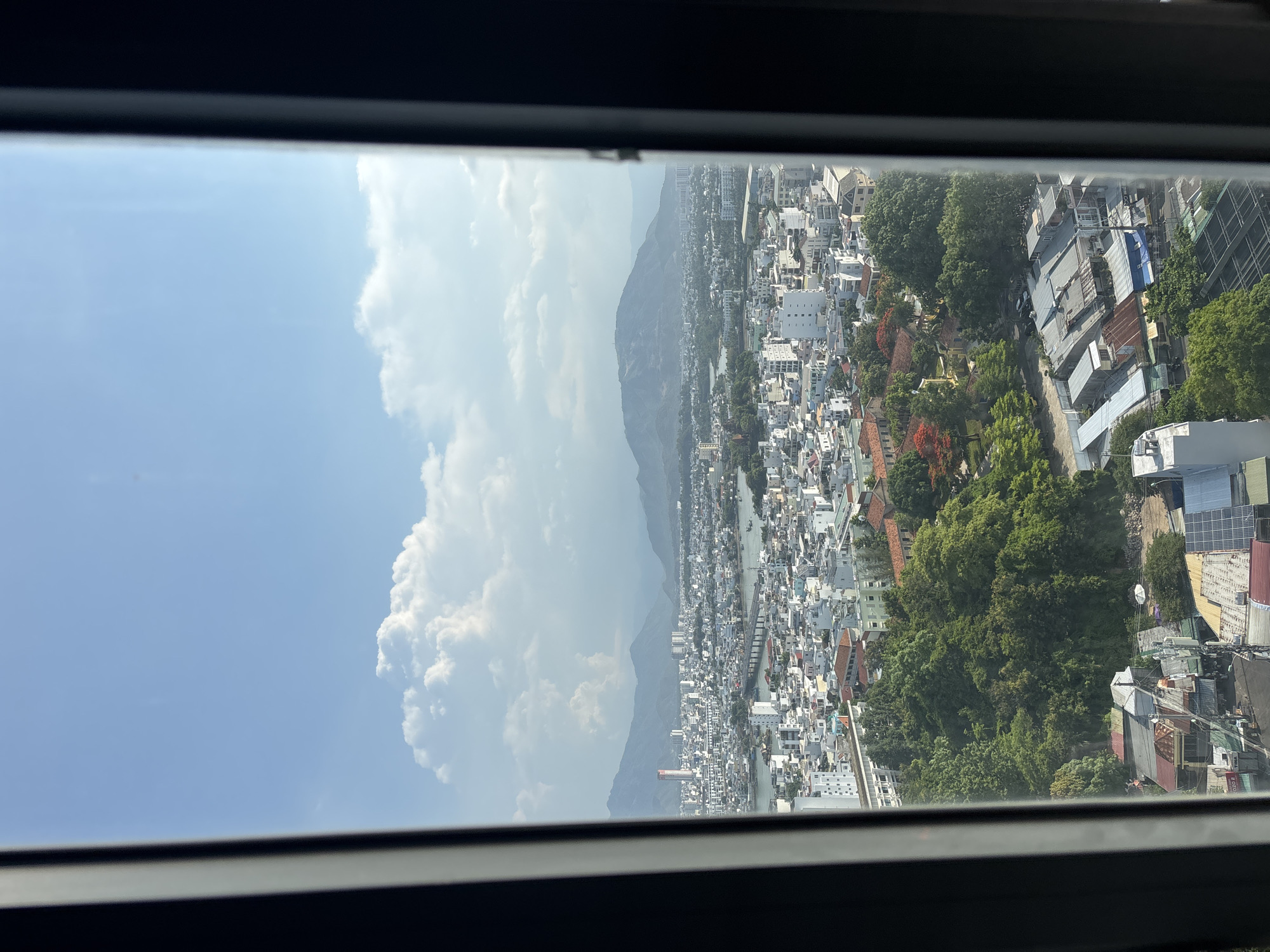 The image is a panoramic view of a coastal city, seen from a high vantage point, possibly a building, framed by dark window frames on the left and right. The foreground features a densely packed urban landscape with a mix of white and pastel-colored buildings, many with flat roofs. Green trees and patches of foliage are interspersed throughout the city, with some larger clusters of trees creating a sense of parkland or natural areas. In the mid-ground, a body of water, likely a river or inlet, runs through the city, crossed by a bridge. Further in the distance, rolling hills and mountains form the backdrop, their slopes covered in a hazy, light green. The sky above is a clear blue, with scattered white cumulus clouds, illuminated by bright sunlight, suggesting a clear, daytime weather condition. A tall, slender building with a red top is visible in the mid-distance of the city. The image appears to be a photograph taken from a window overlooking the urban expanse and natural surroundings.