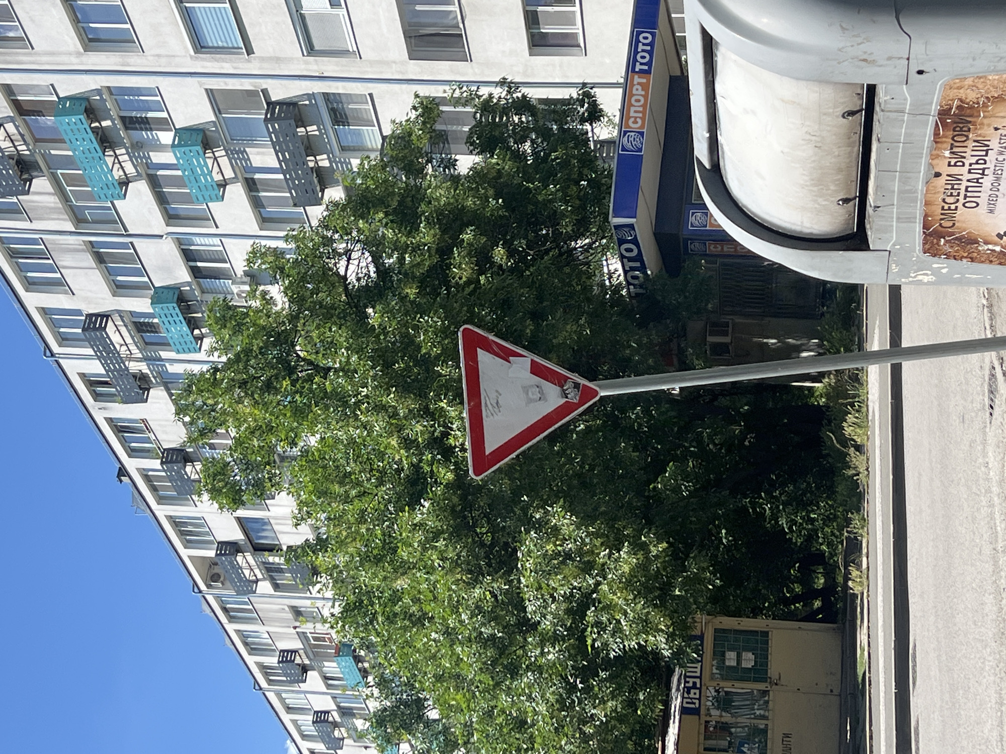 The image shows a street scene in Burgas, Bulgaria. A tall, grey apartment building with rows of windows and small metal balconies occupies the left side of the frame, reaching towards a blue sky. A large, green tree dominates the central portion of the image, partially obscuring a triangular yield sign with a red border. To the right, a "Спорт Тото" (Sport Toto) advertisement is visible. Further right is a grey trash receptacle. A sidewalk and road run along the right edge of the frame. The environment is urban.
