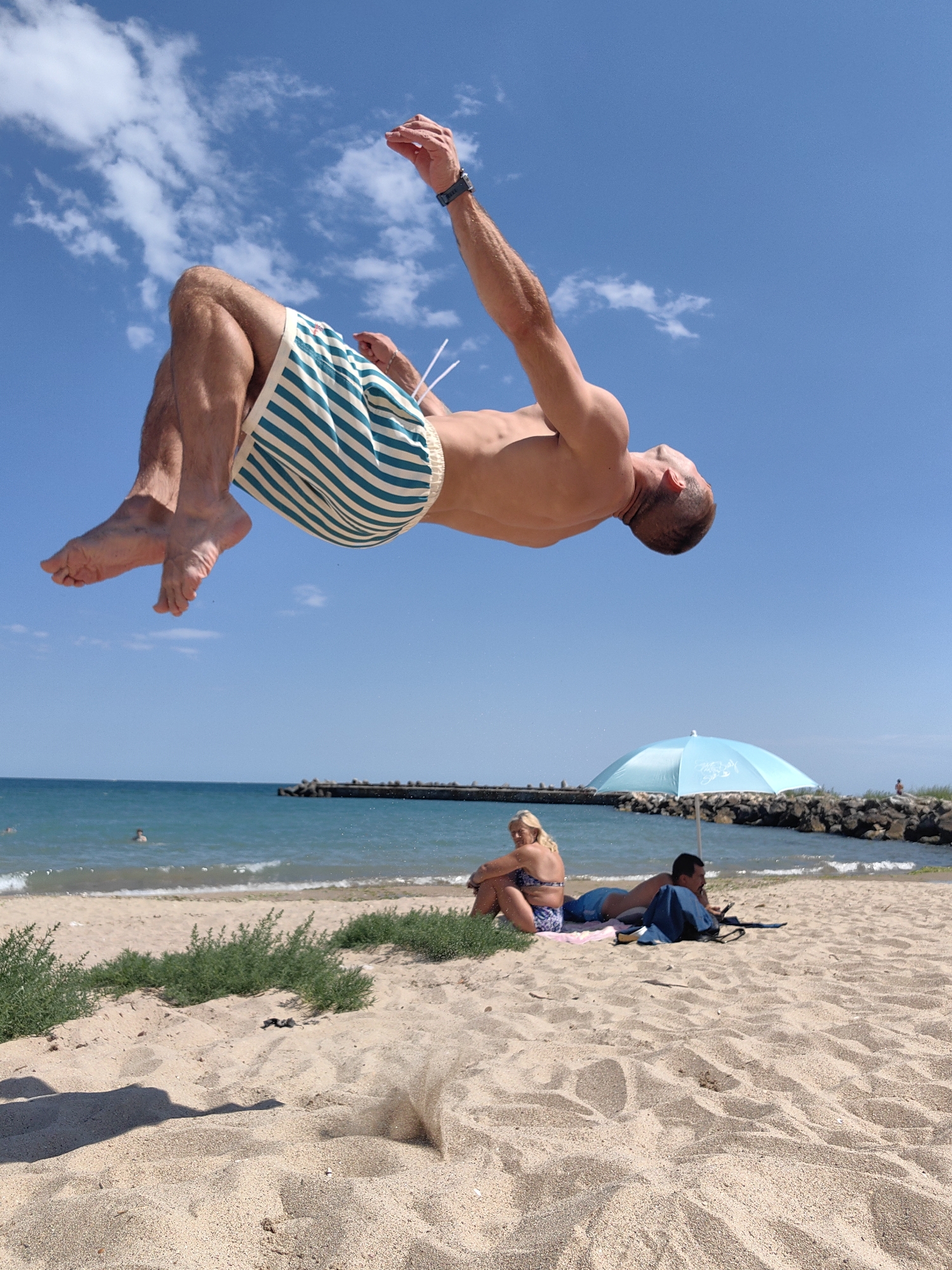 An athletic man is captured mid-flip in the air above a sandy beach under a clear blue sky with scattered clouds. He is wearing blue and white striped swim trunks and a black watch on his left wrist. In the background, the turquoise waters of the Black Sea are visible, with a distant breakwater or pier. On the beach, a woman in a blue and white patterned bikini sits on a pink towel, and a man in blue shorts lies on a towel next to her, with a blue backpack nearby. A light blue umbrella provides shade on the sand. The scene appears to be a public beach, possibly near a coastal city.