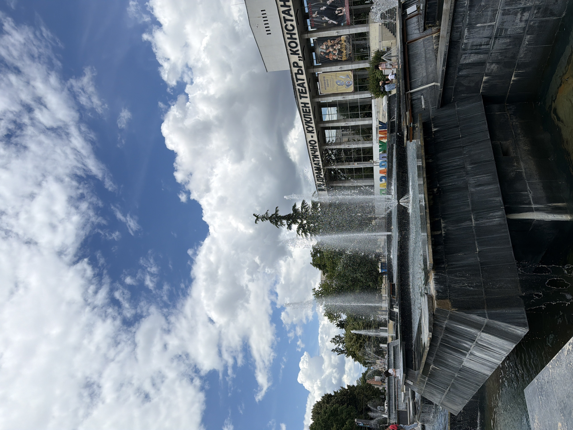 The image depicts the central square of Pazardzhik, Bulgaria, on a cloudy day. In the foreground, a multi-tiered fountain with dark stone cascading elements is active, with water spraying upwards and flowing over the edges. Behind the fountain, a modern building with large windows and signage reads "ДРАМАТИЧНО-КУКЛЕН ТЕАТЪР „КОНСТАНТИН КИРИЛОВ”" which translates to "Drama and Puppet Theatre "Konstantin Kirilov"". The building displays several theatrical posters on its facade. Several people are visible, some standing near the fountain and others on the steps of the theatre. Green trees line the background, and a clear blue sky with prominent white clouds fills the upper portion of the frame.