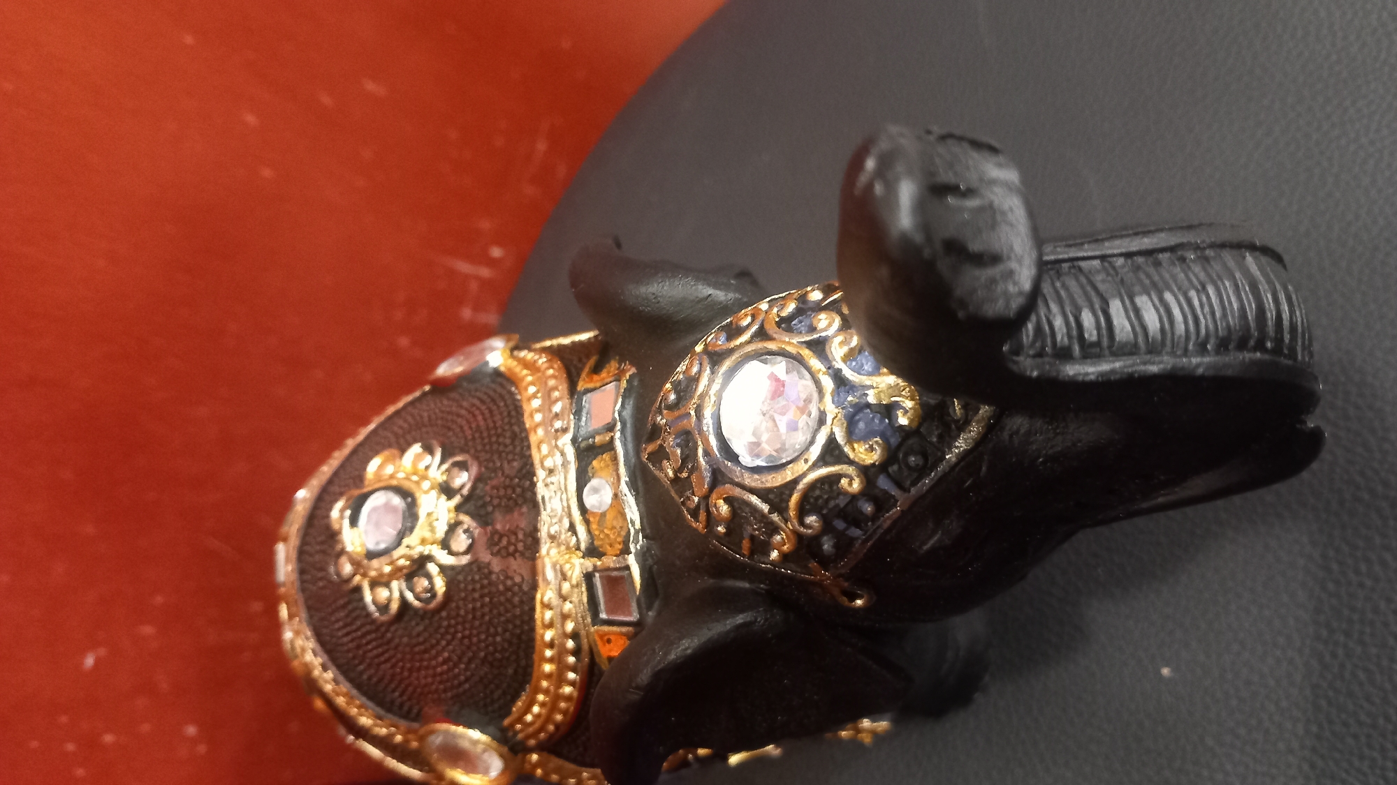 A close-up, shallow depth-of-field, tilted shot captures the head of a decorative black elephant figurine. The figurine is adorned with gold trim, blue accents, and clear, faceted gemstones on its forehead and brow. The elephant is positioned on a dark, textured surface, possibly leather or faux leather. The background is a solid, reddish-orange color with some slight texture and dust specks visible. There are no people or other objects clearly identifiable in the scene. The specific location in Kos, Greece is not determinable from the image content.