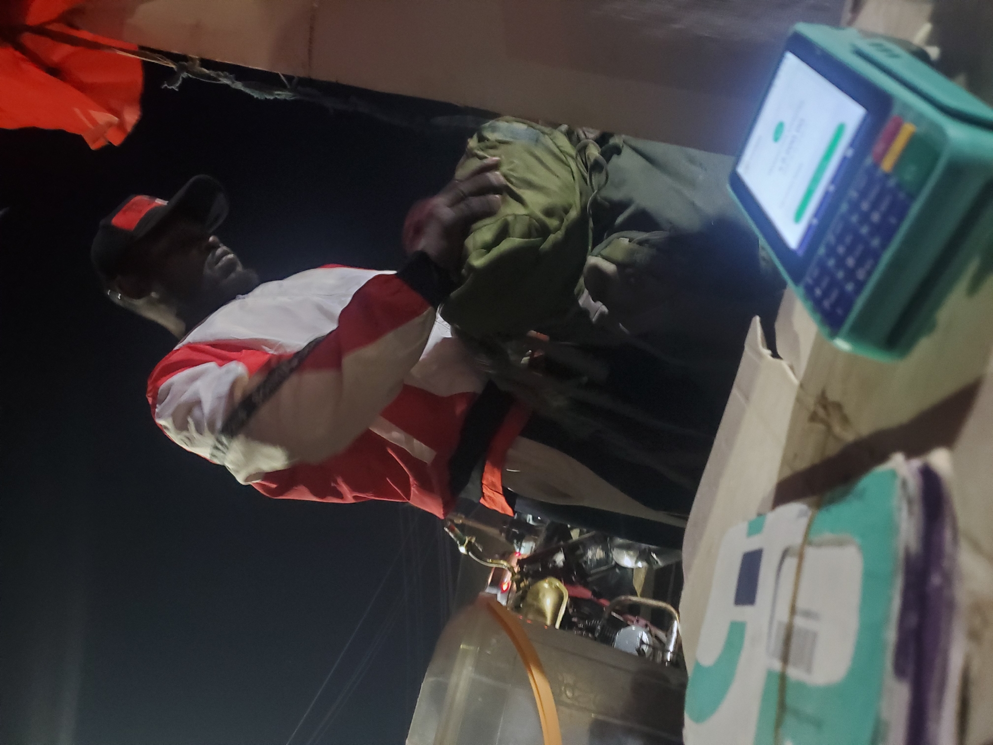 In Gwong, Nigeria, a man wearing a red and white jacket and a black baseball cap with a red logo is standing at a counter. He is holding a green bag and appears to be conducting a transaction at a mobile payment terminal. The terminal is green and displays a white screen with green and blue icons. To the left of the man, a motorcycle is visible, and to the far left, a transparent container with an orange strap is present. The scene is dimly lit, suggesting it is nighttime.