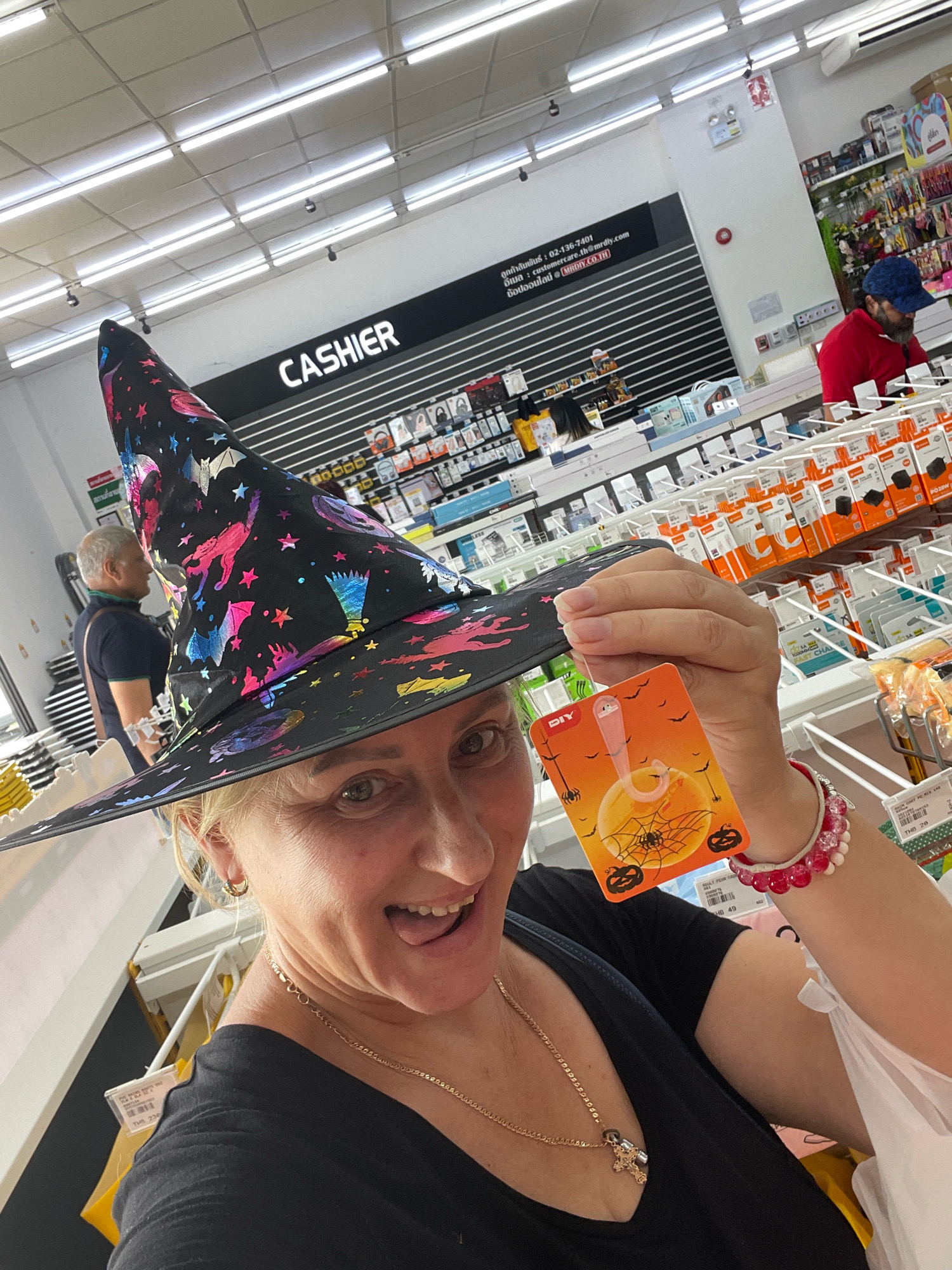 A woman wearing a black witch hat with colorful celestial patterns, a black v-neck shirt, and a gold chain necklace is holding a small orange tag in her right hand. The tag features a spiderweb design and Halloween-themed illustrations. She is indoors, in what appears to be a retail store, with fluorescent lighting and shelves stocked with merchandise in the background. A sign that reads "CASHIER" is visible on a black wall behind her. Another person, a man wearing a red shirt and blue hat, is visible in the background, partially obscured by shelves. The location is identified as a DIY store in Pattaya City, Thailand.