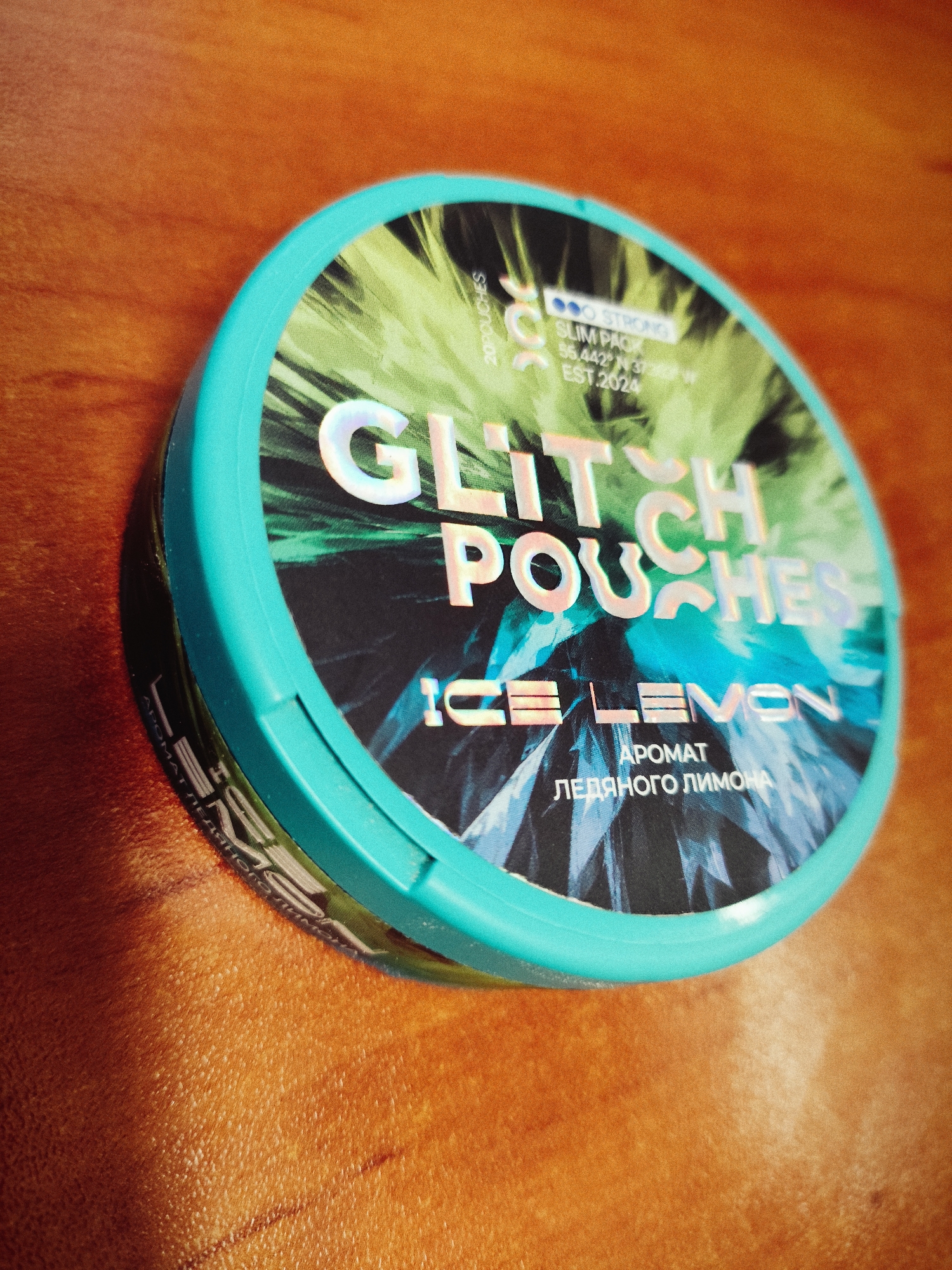 A teal-colored, circular tin of "Glitch Pouches Ice Lemon" sits on a wooden surface. The tin has a holographic design with the brand name prominently displayed in large, iridescent letters. Below the brand name, "ICE LEMON" is written in smaller, holographic letters, followed by Russian text indicating "AROMAT" (flavor) and "LEDYANOGO LIMONA" (icy lemon). The wooden surface appears to be a table or desk, with visible grain and some minor scratches. The lighting suggests an indoor environment, possibly with overhead lighting casting subtle shadows. No people are visible in the image.