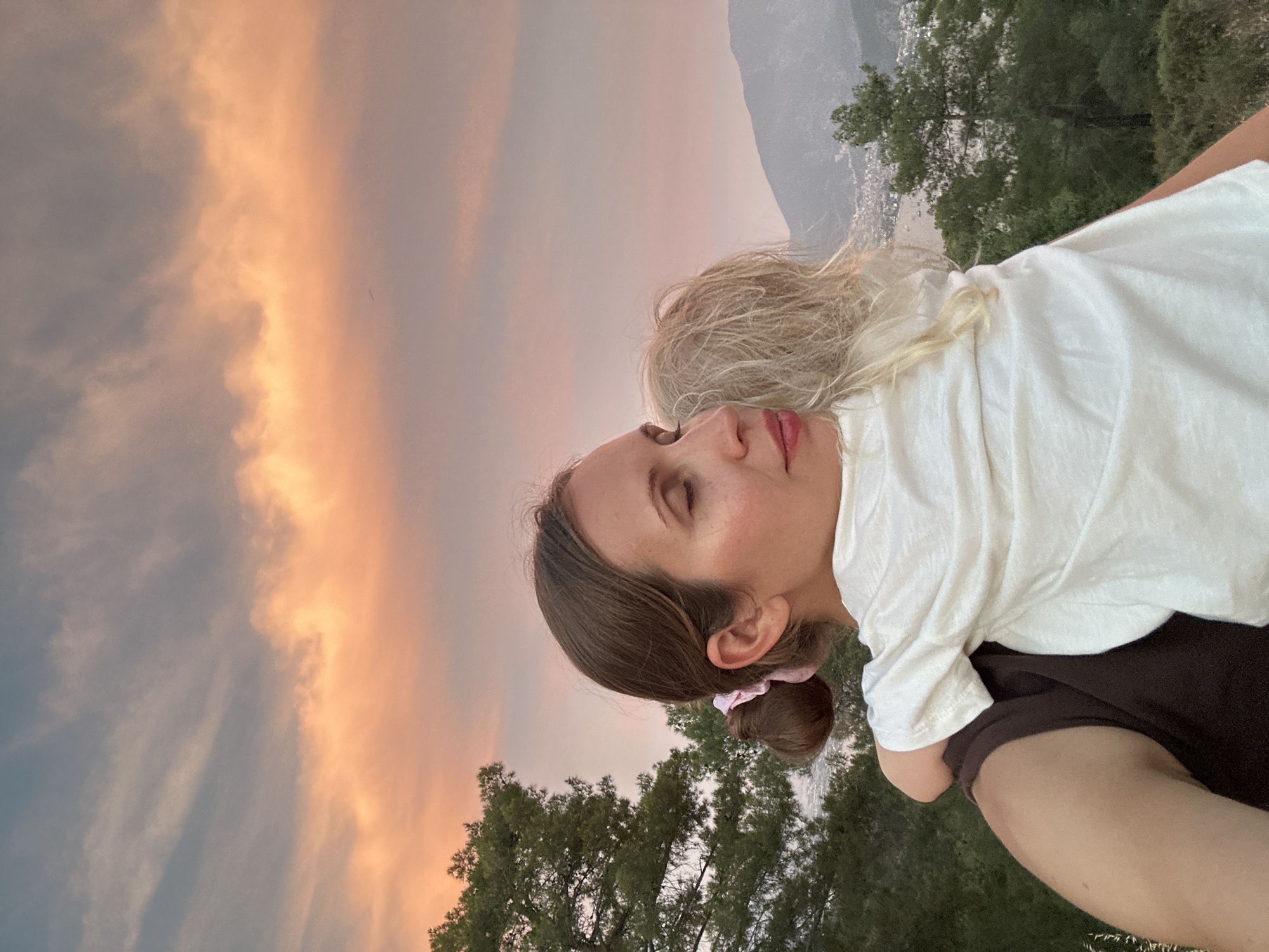 A medium shot captures a woman holding a child outdoors at sunset, with a hint of the city of Fethiye, Turkey, visible in the background. The woman, with her eyes closed and a serene expression, is facing right and embracing the child, who is facing away from the camera. The child has blonde hair and is wearing a white t-shirt. The woman's hair is pulled back into a bun with a pink scrunchie. Behind them, the sky is filled with the warm hues of sunset, casting a soft glow on the scene. Green trees frame the left side of the image, and mountains and a body of water, likely the Mediterranean Sea with a bay and boats, are visible in the distance, suggesting a coastal location near Fethiye. The overall atmosphere is one of tenderness and peace, with the natural beauty of the landscape enhancing the intimate moment between the two individuals.