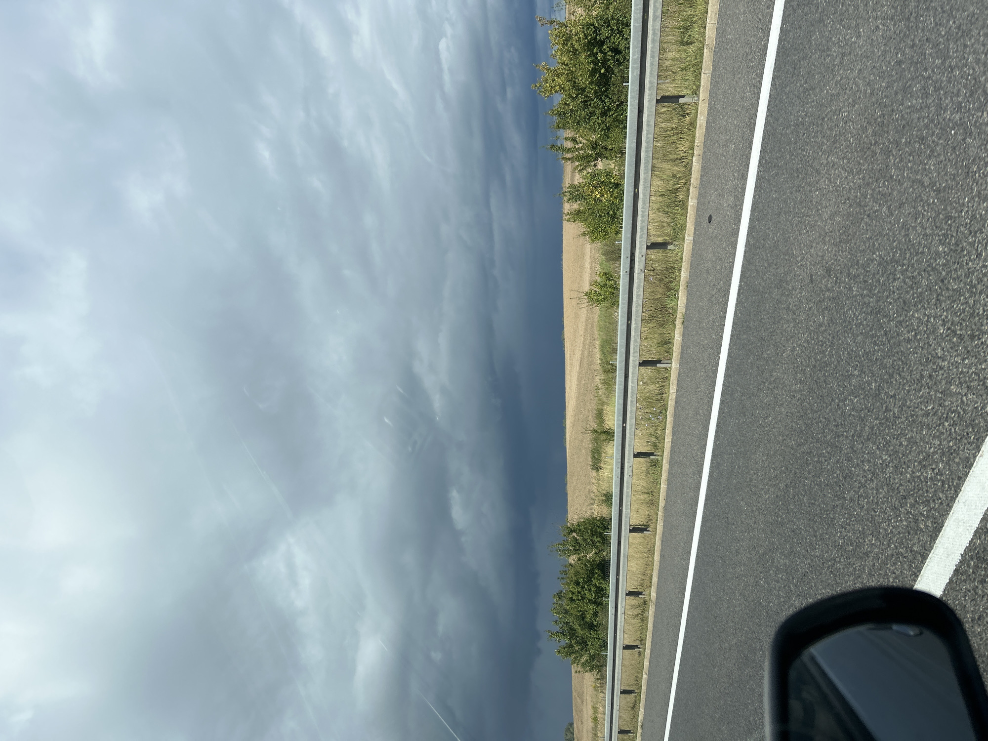 A highway scene in Gościkowo, Poland, under a dramatic, dark, stormy sky. The foreground shows the asphalt road with a white lane marker. To the left, a car's side mirror is visible. Beyond the road is a metal guardrail separating it from a grassy verge. Further back, a field of dry, golden crops stretches to the horizon, bordered by a line of green trees and bushes. The sky is filled with heavy, dark grey clouds, suggesting imminent rain. Faint white contrails from aircraft are visible against the clouds. There are no people or animals visible in the image.