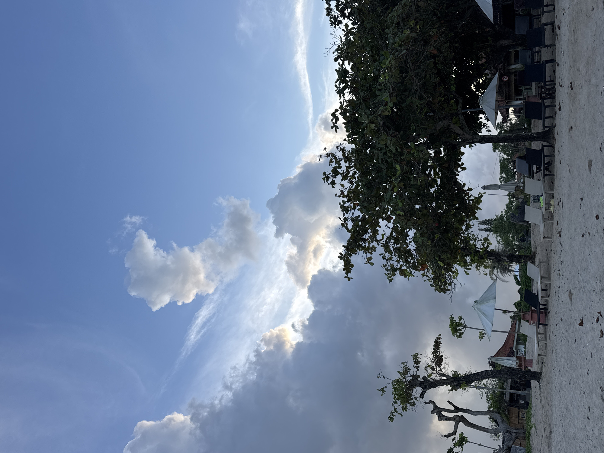 The image captures a daytime scene at Jimbaran, Indonesia, featuring a beachside establishment with a view of the sky. The lower portion of the image displays beach sand, interspersed with trees, chairs, tables, and umbrellas. The upper portion showcases a bright blue sky with varying cloud formations, ranging from wispy cirrus to cumulus clouds. The lighting suggests early afternoon. No people are visible in the frame. The overall mood is serene and relaxed, suggesting a typical day at a beach resort in Jimbaran.