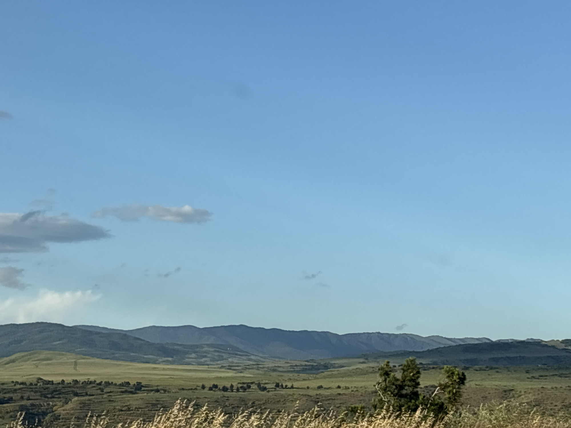 This image depicts a scenic landscape with rolling hills and mountains under a clear blue sky. The foreground is dominated by dry, tall grass, suggesting a dry season or arid environment. In the middle ground, there are green, undulating hills dotted with sparse trees and shrubs. Further in the background, several layers of mountains recede into the distance, their peaks softened by atmospheric haze. A few wispy clouds are scattered across the sky. The overall impression is one of a vast, open natural setting. The lighting suggests it could be late afternoon or early morning, with the sun casting a gentle light on the landscape. There is no visible text in the image. The scene evokes a sense of tranquility and the grandeur of nature.