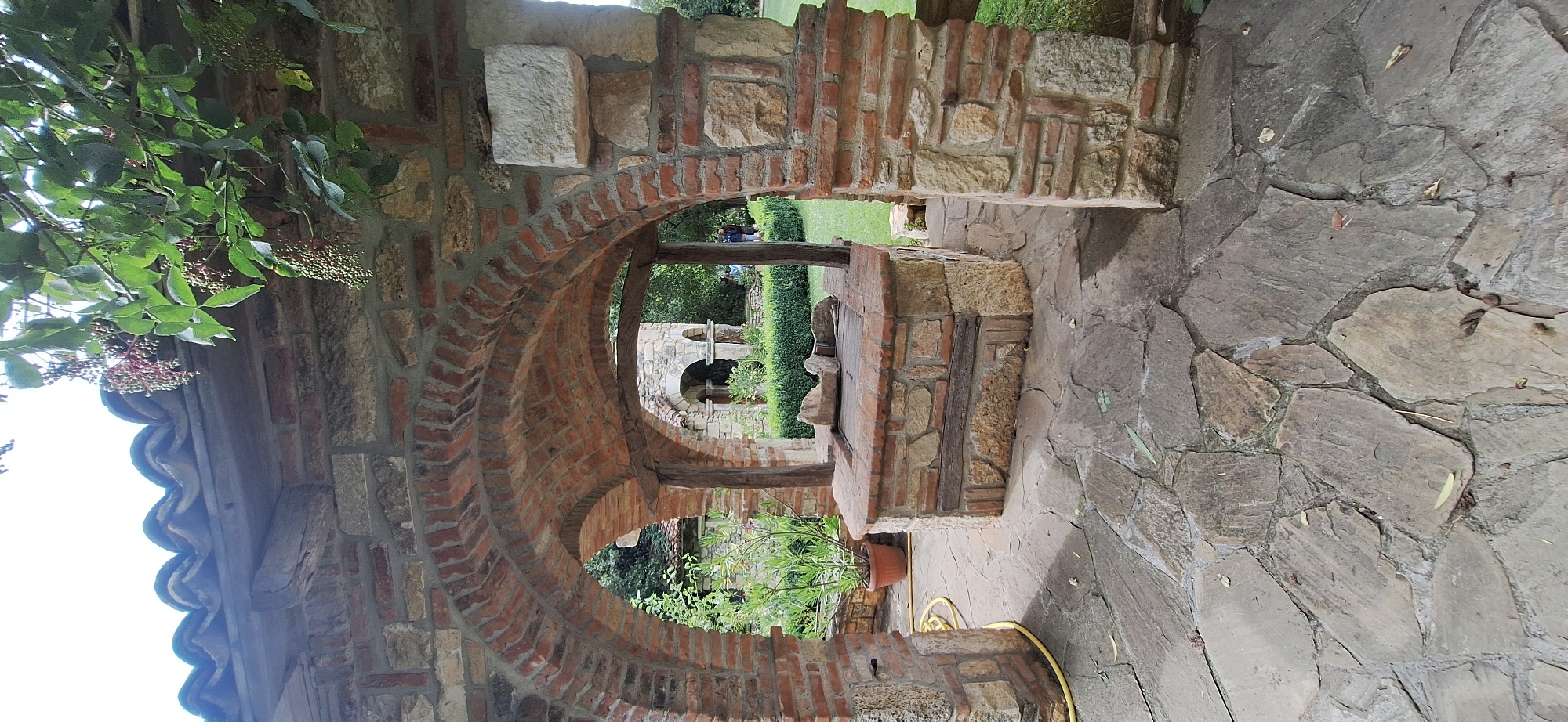 The image shows an arched structure built with red bricks and stone blocks, likely part of a historical site in Kalimantsi, Bulgaria. In the foreground, a stone-paved courtyard leads to a well with a brick and stone structure. The well has a wooden plank cover and appears to be made of layered stone and brick. A terracotta pot with a plant is visible to the left of the well, next to a coiled yellow hose. Behind the well, through the archway, a garden area with trimmed hedges and a building with similar architectural elements can be seen. Two figures, appearing to be adults, are walking in the background garden. Overhanging the structure is a tree with green leaves and clusters of small, reddish-pink berries. The sky is visible through the archway and above the structure. The overall scene depicts an outdoor historical or cultural setting.