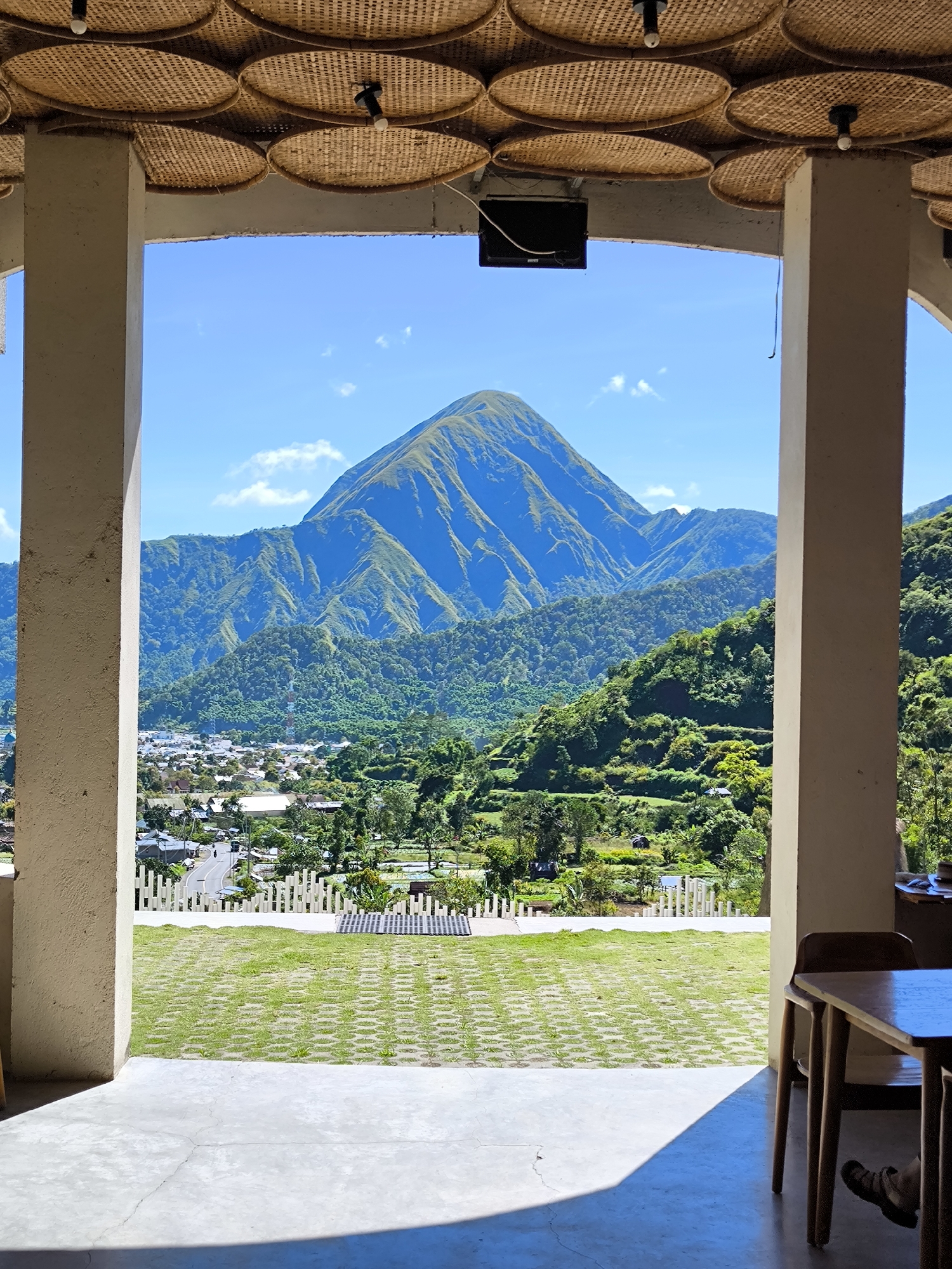 The image shows a view framed by two pillars, likely from a building, overlooking Sembalun Bumbung, Indonesia. The landscape includes a prominent mountain, lush green hills with terraced agriculture, and a town with houses and roads. A grassy area with patterned paving is visible in the foreground. Above, the ceiling has a decorative woven pattern with interspersed lights. A black electronic device is mounted on the ceiling between the pillars. A table and chairs are partially visible on the right side of the frame. The sky is blue with some clouds. The scene appears to be taken during daylight hours.