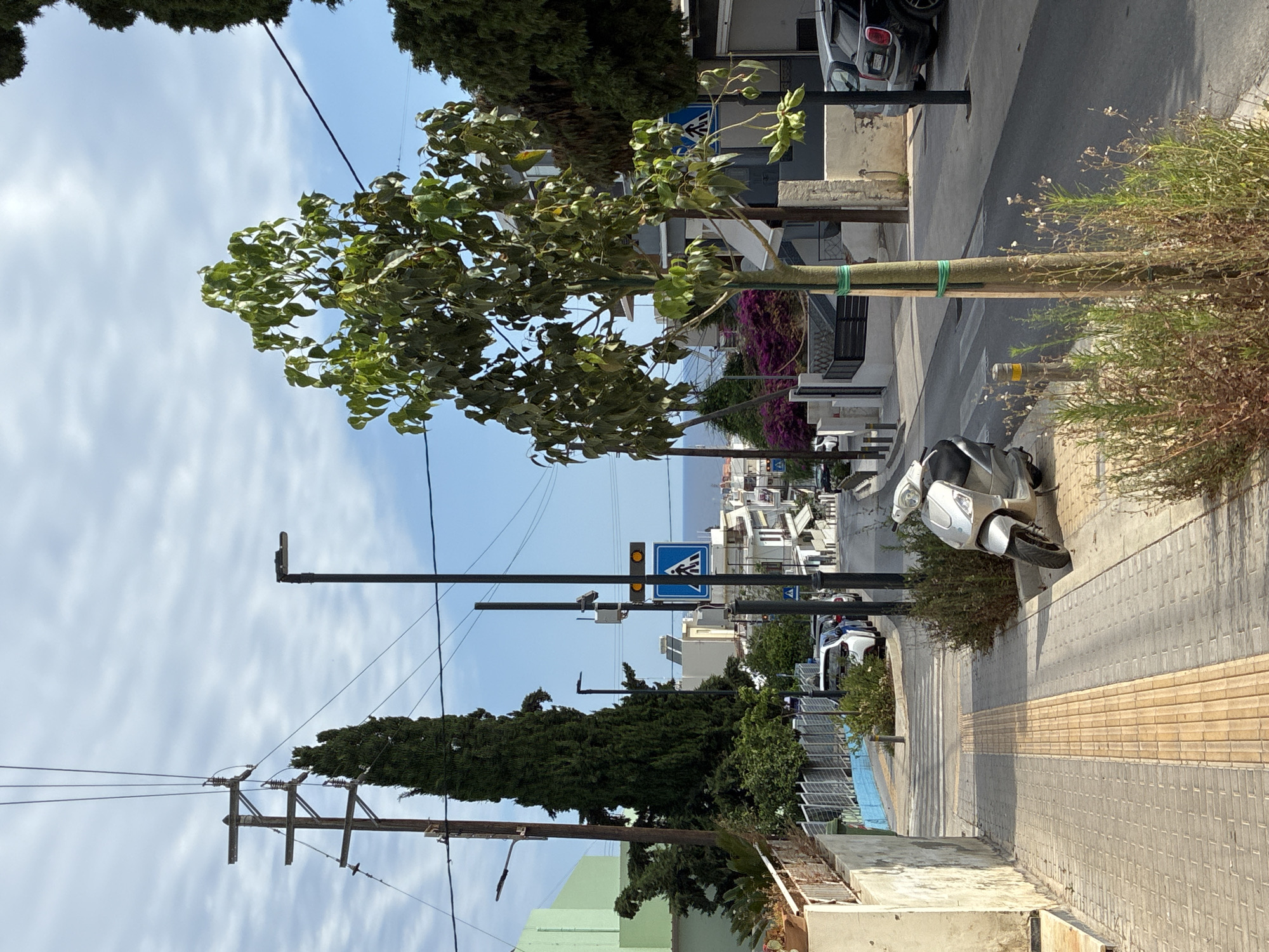 The image depicts a street scene in Rethymno, Greece. A silver scooter is parked on the right side of a pedestrian walkway. The walkway is paved with grey bricks and features a tactile paving strip for visually impaired pedestrians. A young tree, supported by green ties, stands in a patch of vegetation on the edge of the walkway.
The street slopes downwards, with buildings lining the right side. These buildings are a mix of styles, some with white facades and balconies, others with darker grey exteriors. A few cars are parked along the street, including a black SUV on the far right. On the left side of the image, there is a wall and a light green building.
In the background, a pedestrian crossing is visible, marked by white stripes on the asphalt. A pedestrian crossing sign with a blue background and a white silhouette of a person walking is also present, along with a traffic light showing two amber lights. Further down the street, the sea is visible under a blue sky with scattered white clouds. Overhead, power lines crisscross the scene, supported by wooden and metal poles. Several tall, slender evergreen trees are also present in the landscape, adding to the Mediterranean feel of the location.