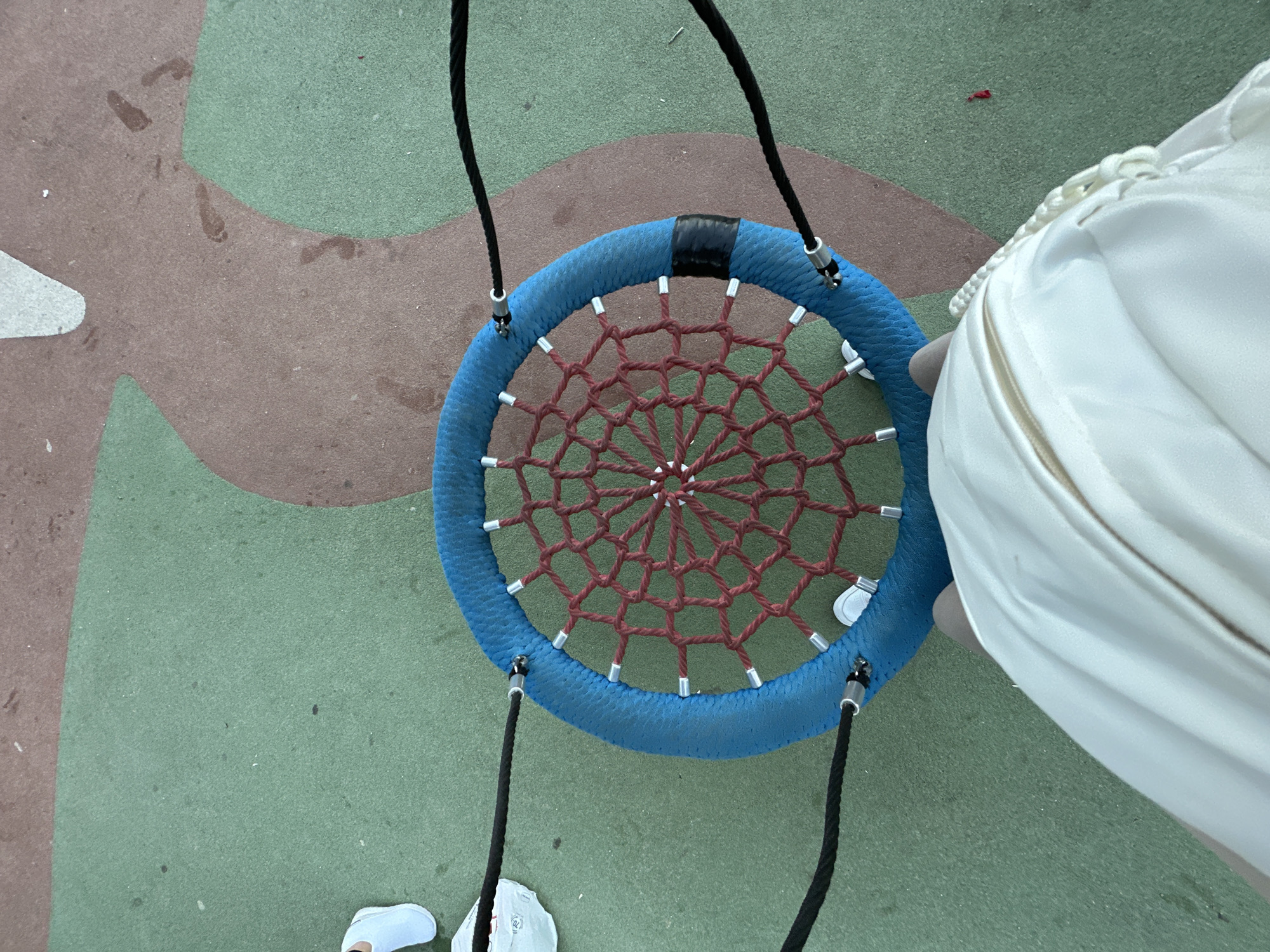 An overhead shot shows a person's legs and torso, clad in white, standing on a colorful playground surface. The person is wearing white sneakers. In front of the person is a large, circular blue swing with a red webbed net in the center. Black ropes suspend the swing. The playground surface is a mosaic of green and brown sections, with some faint footprints visible on the brown area. The scene appears to be in a public park or playground area in Sant Feliu de Guíxols, Spain.