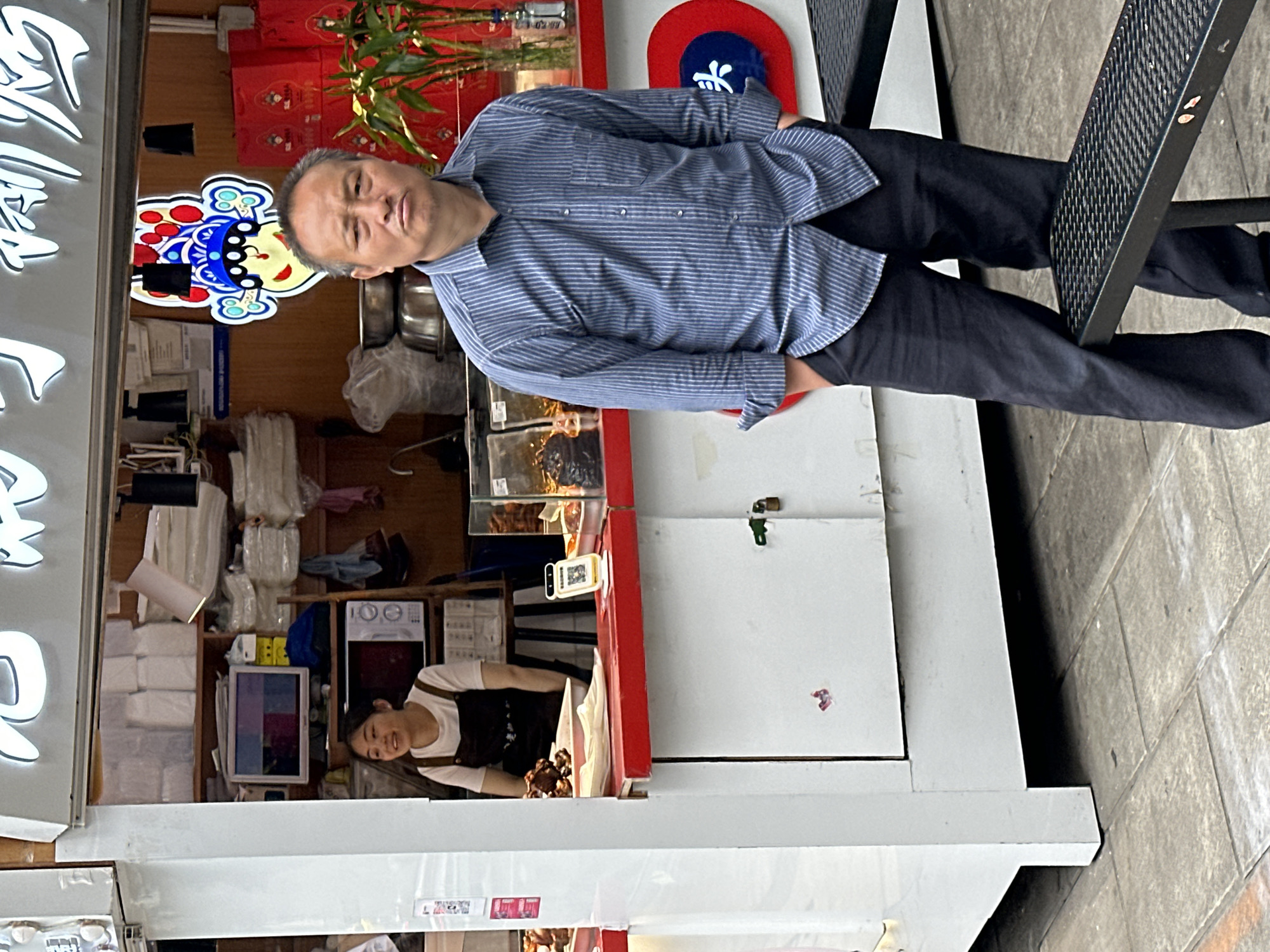 The image captures a street scene in what appears to be the Yuzhong District of China, based on architectural and cultural cues. A middle-aged man stands to the right, wearing a striped shirt and dark pants, partially obscured by a metal outdoor chair. The man appears to be looking at the camera. Behind him is a storefront with Chinese signage. Inside the storefront, a woman in a dark apron is visible behind a counter, likely serving customers. The store contains various items, including packaged goods, equipment and what appears to be food in a display case. The overall scene conveys a snapshot of daily life in a bustling urban environment.