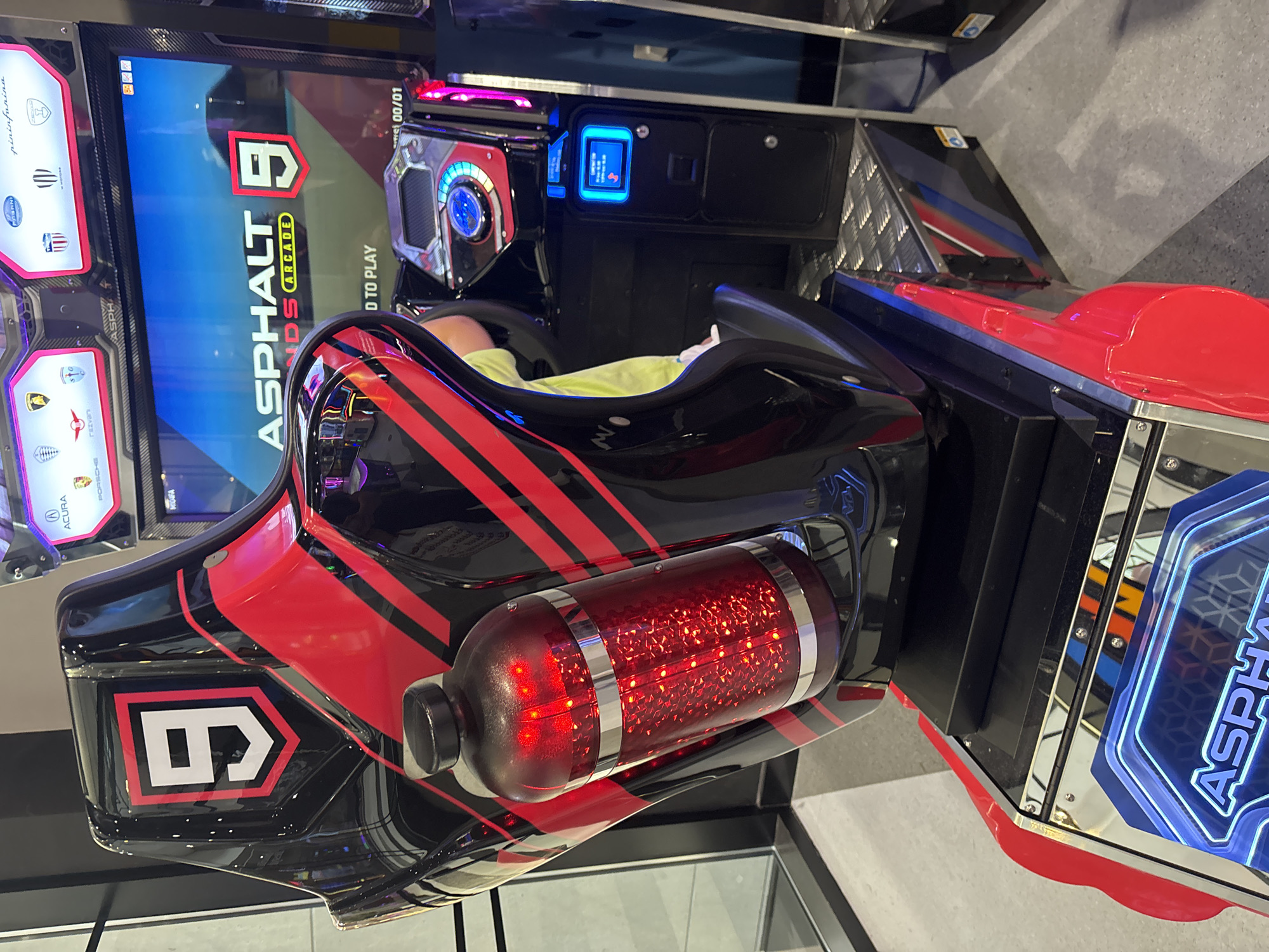 This image shows an arcade racing game machine, specifically for "Asphalt 9: Legends Arcade," at an indoor entertainment venue in Dubai, United Arab Emirates. The machine features a highly stylized, black and red racing seat designed to immerse the player. A prominent "9" in a white shield shape with a red border is visible on the backrest of the seat. A red, illuminated cylinder, possibly a visual effect or accessory, is attached to the side of the seat. The game's title, "ASPHALT 9: LEGENDS ARCADE," is displayed on the large screen at the top of the machine, along with the prompt "PRESS START TO PLAY." The screen also shows various car manufacturer logos. Below the screen, various control panels and a touch screen interface are visible. The floor of the venue is a textured metal grate. The lighting is a mix of ambient overhead lighting and the colorful neon lights emanating from the arcade machines. No individuals are clearly visible interacting with this specific machine, though other arcade machines and people may be present in the background, suggested by reflections and the general ambiance of an arcade. The overall impression is of a modern, high-tech entertainment setting.