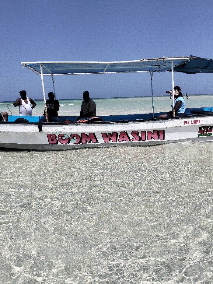 A white, flat-bottomed boat named 'BOOM WASINI' rests in shallow, clear water in Wasini, Kenya. The boat's starboard side displays 'BOOM WASINI' in pink letters, a Kenyan flag decal, and the text 'NI LIPI'. A light blue fabric canopy is supported by a metal frame above the boat's interior. Four individuals are present: one male in a white sleeveless top stands at the forward-left, two males are seated mid-boat (one in a dark t-shirt, the other in a light long-sleeved shirt), and one female in a light blue short-sleeved top and striped visor is seated at the mid-right, holding an object to her right eye. The water around the boat is transparent, revealing the sandy seabed, and extends to a light turquoise hue at the distant horizon under a clear blue sky. Faint human figures are discernible on the water's surface in the background.