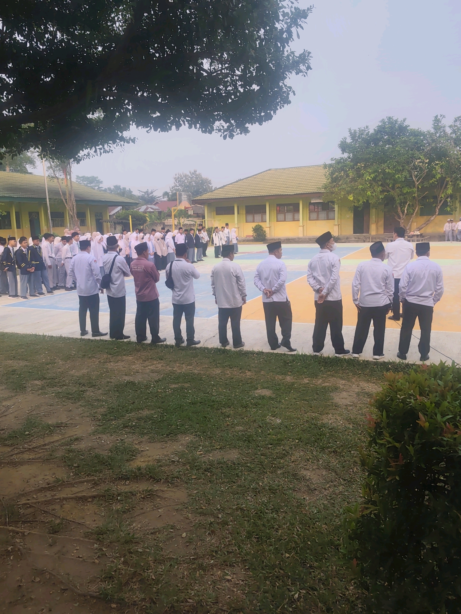 A group of approximately 30 to 40 individuals, primarily young people, are assembled in an outdoor courtyard. The individuals are dressed in various forms of attire, including school uniforms, traditional Islamic clothing, and formal shirts and trousers. The setting appears to be a school or educational institution, evidenced by the presence of a building in the background with a yellowish facade and a green tiled roof. The courtyard is paved with a multi-colored surface, featuring blue, orange, and white sections, possibly indicating a sports court. A large tree with dense foliage is visible in the foreground, casting shadows. The sky is overcast, suggesting a cloudy day. Some individuals are standing in neat rows, while others are gathered in smaller groups. The overall atmosphere suggests an organized event or gathering. The location is identified as Pematang Reba, Indonesia.