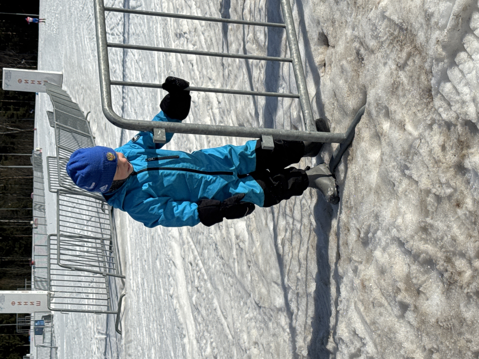 An outdoor scene in Прикамский, Russia, depicts a young child standing in a snowy environment, likely a winter sports venue. The child, dressed in a bright blue snowsuit, a blue knitted hat with a gold double-headed eagle emblem, black gloves, and dark waterproof boots, is positioned in the center-right of the frame, looking downwards. Both gloved hands are grasping a vertical bar of a gray metal crowd control barrier that extends across the midground. The foreground consists of uneven, partially melted, and soiled snow showing footprints. Behind the child, multiple similar metal barriers are arranged in rows, indicating a demarcated area or course. In the mid-background, a white vertical sign with red Cyrillic text reading "ФИНИШ" (FINISH) is visible. Further in the background, a dense line of dark evergreen trees borders the snow-covered area under a bright sky. A blurred figure in pink and white winter attire is discernible in the far right background, possibly a skier. Shadows from the child and barriers are cast onto the snow.