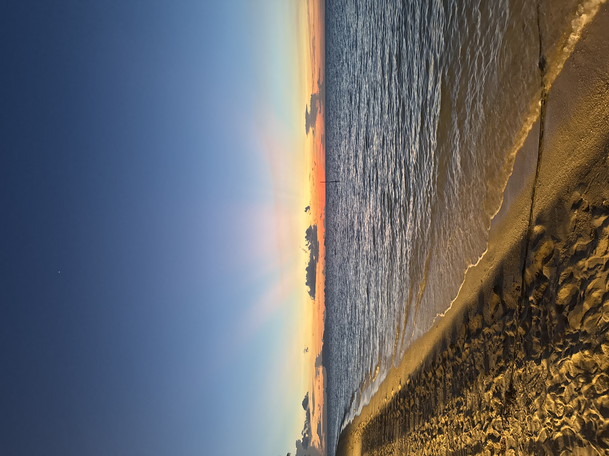 This media file captures a serene beach scene at sunset. The image is oriented vertically, showing a vast expanse of the ocean on the right side, with gentle waves lapping at a sandy shore. The sand is textured, showing ripples and shadows cast by an unseen light source, possibly artificial.
The sky on the left transitions from a deep, clear blue at the top to lighter shades of blue, pink, and orange near the horizon, indicative of a sunset. Rays of sunlight emanate from behind a cluster of dark clouds near the horizon, creating a dramatic and picturesque effect. A thin, dark vertical line, possibly a post or a distant structure, is visible in the water near the horizon line. Two tiny bright specks are present in the deep blue part of the sky, which could be stars or distant lights.
The overall mood of the image is peaceful and tranquil, evoking the beauty of nature at the end of the day. There is no visible text in the image. The time of day is clearly sunset, and the weather appears clear with some cloud formations. The location is a beach, but specific geographical cues are not evident. There are no people or distinguishable objects other than the natural elements of the sea, sky, and sand.
