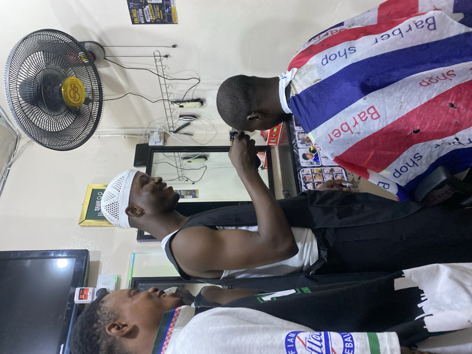 The image shows a barber shop scene, likely in Ado, Nigeria. A man with a white kufi cap is holding a smartphone, possibly taking a photo. Another man sits under a barber cape with red, white, and blue stripes. A third man is in the foreground. Behind them is a wall-mounted fan, electrical outlets, clippers hanging on a rack, a mirror, and a television. A sign reads "EBLINGS MUSIC STUDIO" and "SHAVEOO" is visible. Logos "FIAN" and "tla" are visible on a t-shirt.