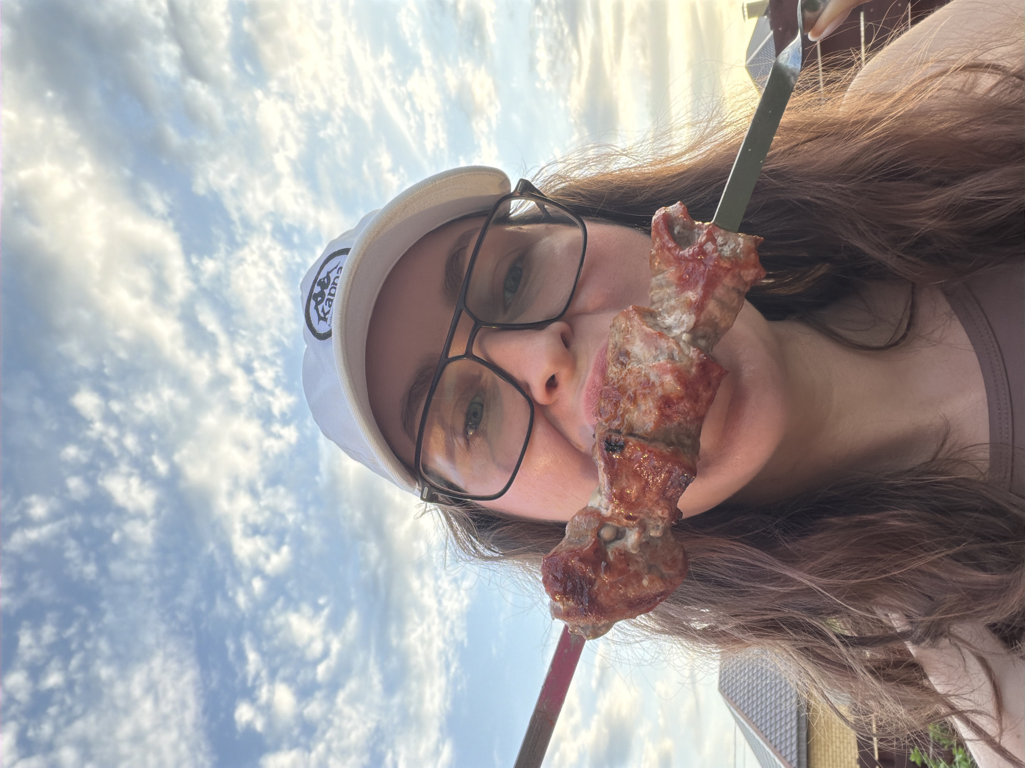 A person, presumably female, is facing the camera, holding a shish kebab skewer up to their mouth. They wear a white baseball cap with a logo, and black-rimmed glasses. The sky is visible in the background, filled with fluffy clouds. The location may be Chaikovsky, Russia, as requested.