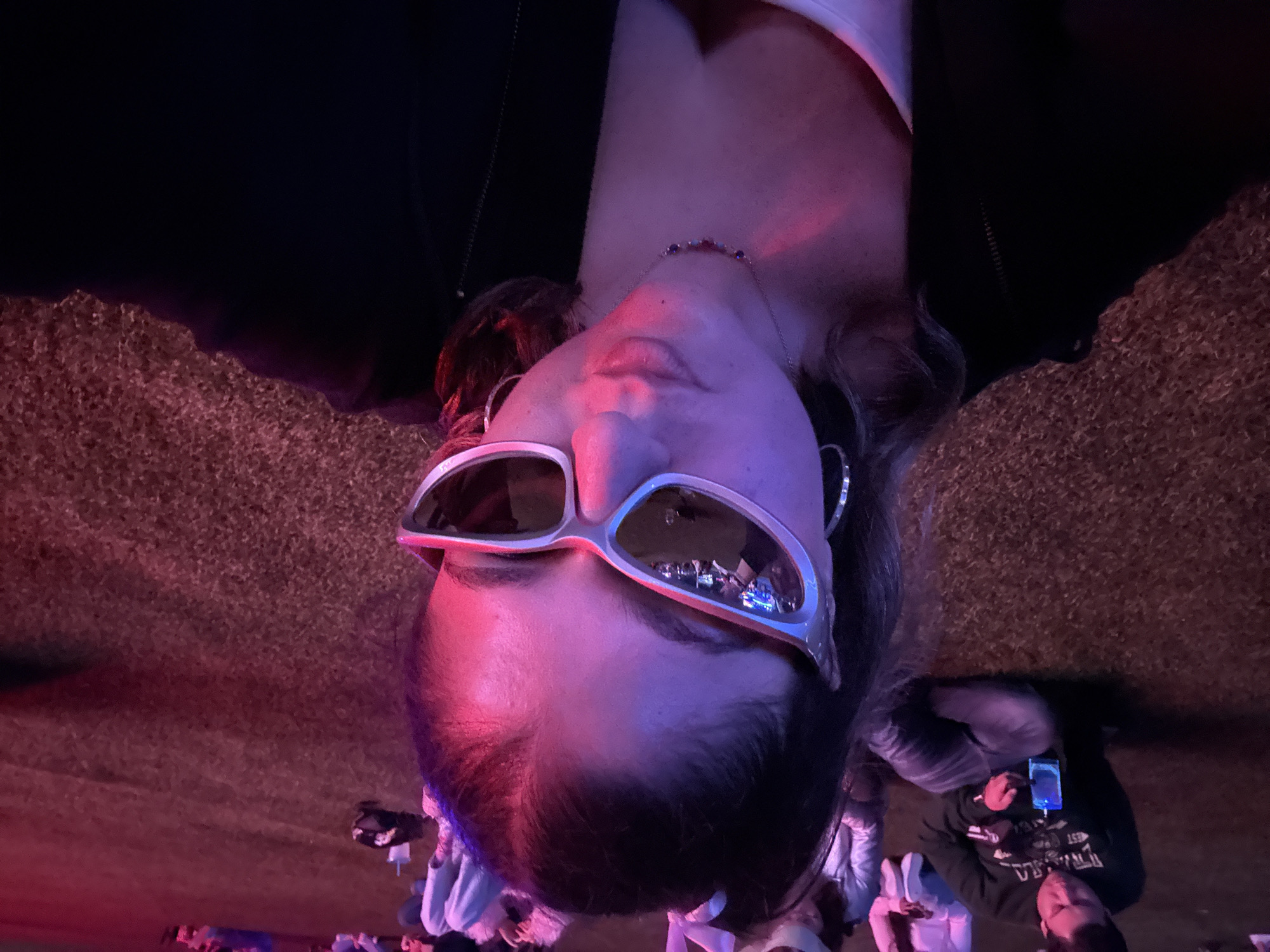 A head-on, upside-down shot shows a person wearing sunglasses with a silver frame and a necklace. The sunglasses reflect the scene they are in, appearing to be in Mexico City. The person is surrounded by other people, some looking at their phones. The background appears to be a field or grassy area. The lighting is dim with a purple hue.