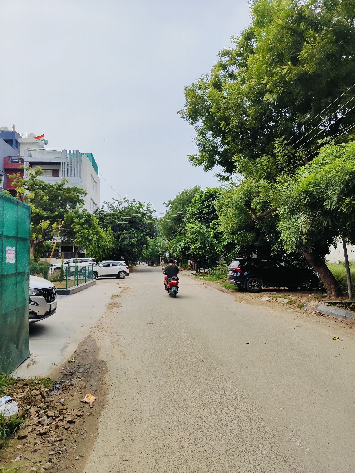 A wide, paved road runs through a residential area in Gurgaon, India. On the left, a modern apartment building with a blue and white facade is visible, topped by a small Indian flag. Adjacent to the building is a green mesh fence. Several white cars are parked along the side of the road. The road is lined with lush green trees, some of which arch over the asphalt. A black SUV is parked under one of these trees on the right side of the road. In the middle of the road, a person on a red electric scooter is riding away from the camera. The sky is overcast. The area appears to be a well-maintained but somewhat cluttered urban environment with debris, including scattered rocks and a plastic wrapper, visible on the side of the road.