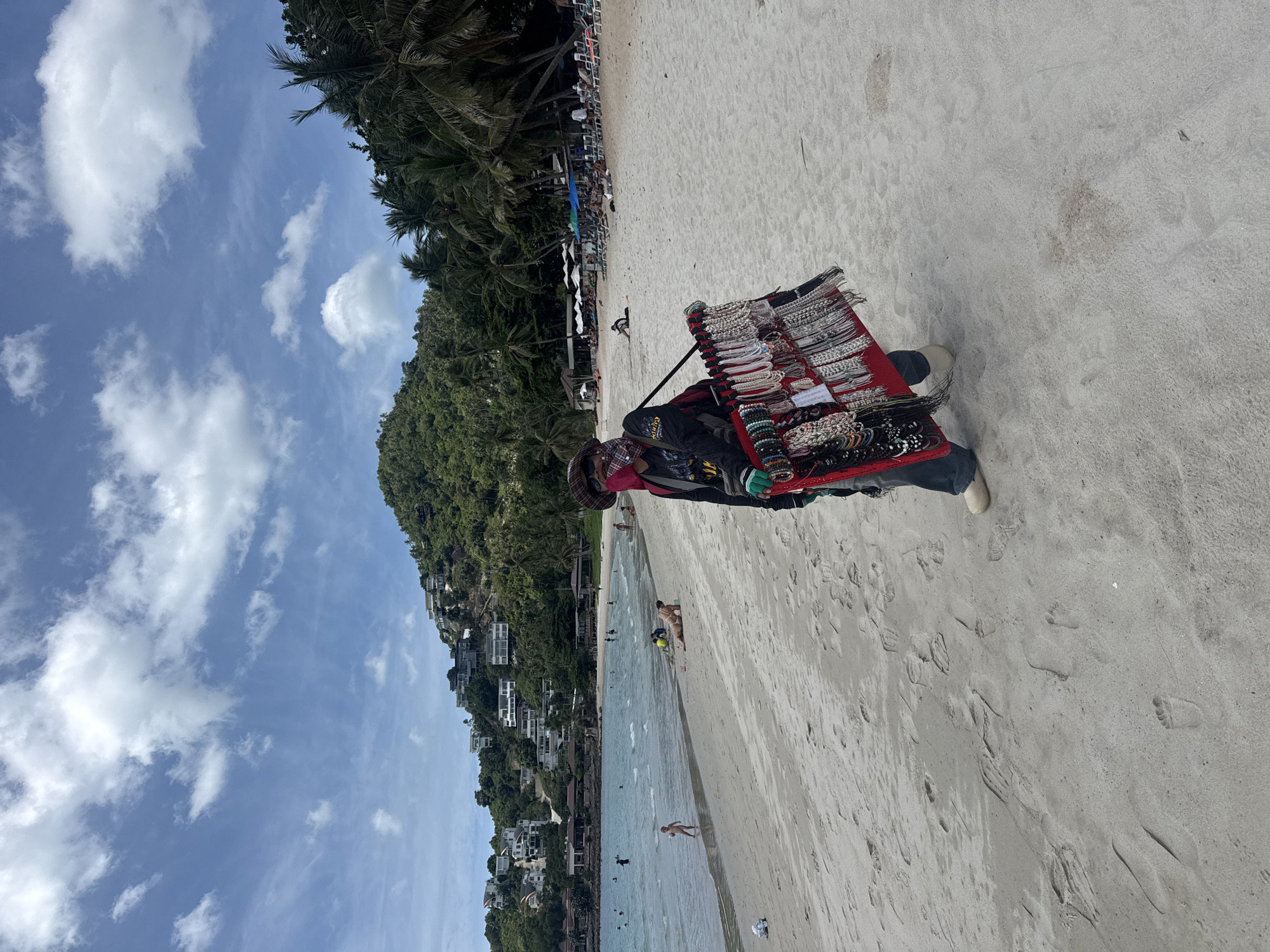 A person stands on a sandy beach in the Ko Samui District of Thailand, facing the ocean. They are wearing a hat, face covering, dark long-sleeved shirt, and jeans, and carrying a display of jewelry around their neck and chest. The jewelry is presented on a red board and consists of various necklaces and bracelets. The beach is populated with a few other individuals, some swimming in the shallow water, and others relaxing on lounge chairs and under umbrellas further down the beach. Palm trees and lush green hills dotted with buildings rise in the background. The sky is blue with scattered white clouds.