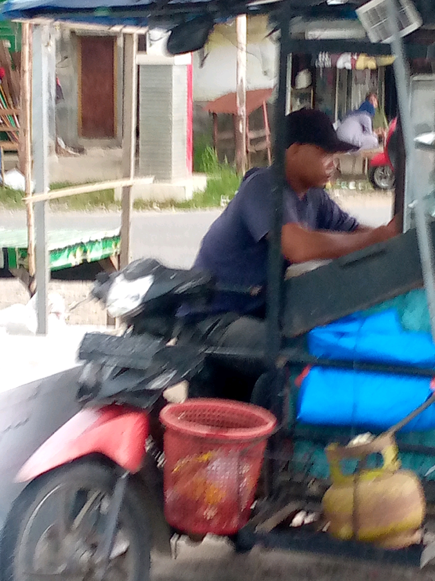 A low-angle, medium shot captures a street scene in Keupoknibong, Indonesia, focusing on a motorized cart. A man wearing a dark baseball cap and a navy blue t-shirt is seated on the cart, his arms resting on the dashboard. He appears to be operating the vehicle. Beside him, a red basket sits on the cart, containing what looks like produce. To the right of the basket, a yellow propane tank is visible. The cart itself is a dark color and is attached to a motorcycle. In the background, a person in a light blue headscarf and a white garment is seated on a bench, looking to the left. Behind them, a structure with a red-trimmed doorway is visible. The environment appears to be a roadside with some greenery and a paved surface.