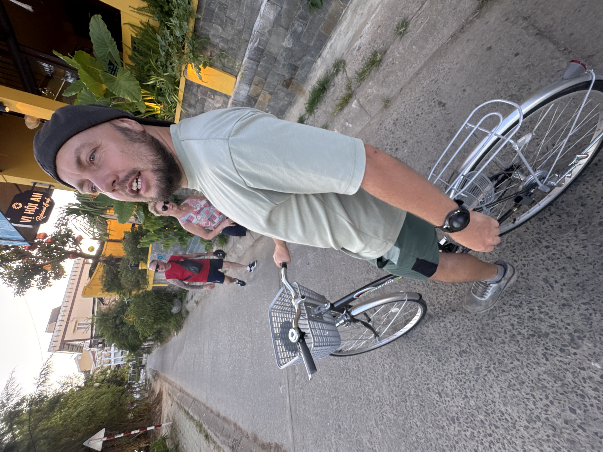 A man wearing a gray t-shirt, green shorts, a black cap, and gray shoes is standing on a street next to a bicycle with a basket on the front. The background includes buildings, trees, and two other people walking in the same direction. The sign for "Vỉ Hội An" is visible. The location is likely Hội An, not Đà Nẵng.