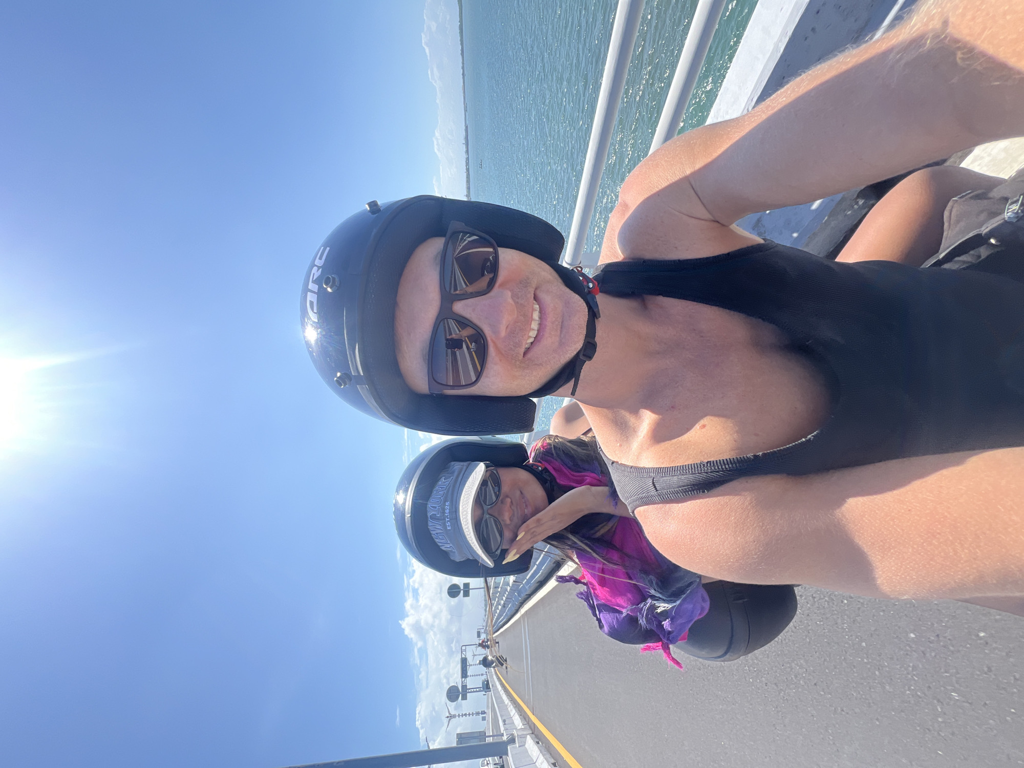 In Kuta Selatan, Indonesia, two individuals wearing helmets and sunglasses are captured in a selfie while riding on a bridge over a body of water. The person in the foreground, on the right, is a fair-skinned male wearing a black tank top. The person behind them, on the left, is a fair-skinned female with a pink and purple scarf around her neck and a gray baseball cap. Both are smiling. The bridge has a white railing and a paved road. The environment is sunny with a bright blue sky and scattered clouds. The water appears calm and blue. Several other people on motorcycles are visible in the background on the bridge, moving away from the camera. The scene suggests a recreational activity, likely a scenic motorcycle ride.