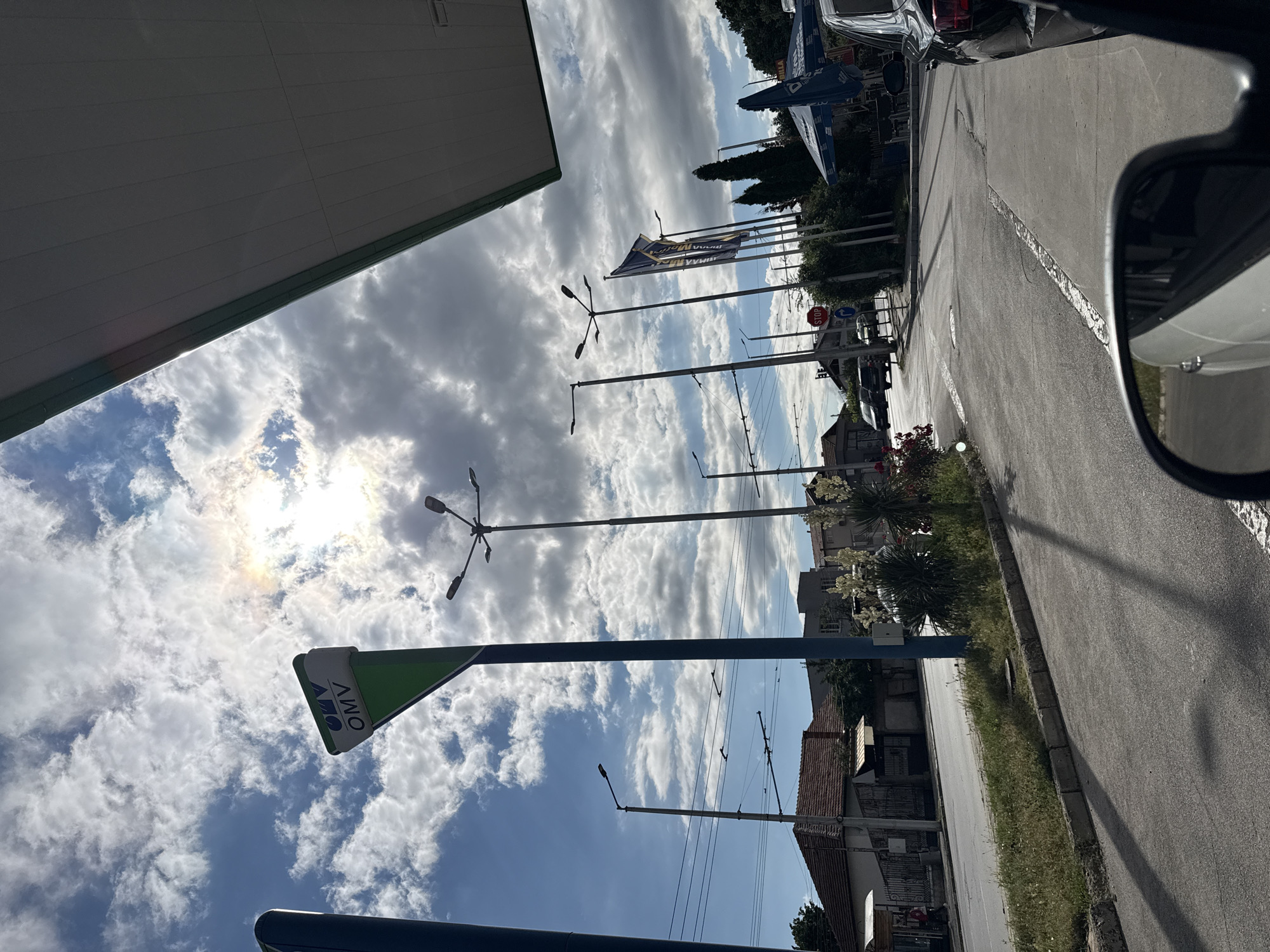 The image captures a street scene in Vratsa, Bulgaria, under a partly cloudy sky. In the foreground, a portion of a car's interior is visible, including a side mirror reflecting a white vehicle. The scene appears to be from inside a vehicle driving or stopped near a gas station.
The dominant elements include a tall blue pole with an OMV sign, triangular in shape with the OMV logo, pointing upwards against the sky. Multiple streetlights are present, some with multiple arms. Overhead wires, possibly for public transport, are strung across the sky. On the left side of the road, residential buildings with red-tiled roofs are visible. Further down the street, several flags are flying, with one prominently displaying blue and yellow colors, and text that appears to be in Cyrillic. A blue umbrella canopy is also visible, suggesting an outdoor seating area or a shop.
The road itself is paved, with painted lane markings. A stop sign is visible on the right side of the road. The overall impression is of a typical urban environment on a day with dynamic cloud formations and sunlight filtering through. The presence of the OMV sign indicates a fuel station, a common feature in urban and roadside settings. The scene suggests a moment captured from a passing vehicle, offering a snapshot of daily life in this Bulgarian city.
