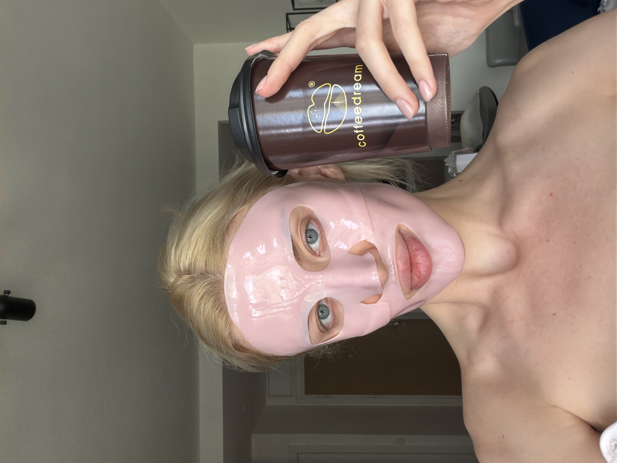 A fair-skinned person is holding a brown and black coffee cup with the logo "coffeedream" over a pink facial mask. The person has blonde hair and blue eyes. The cup is being held in their right hand. The background is a blurred interior scene with light gray walls. The location cannot be determined without more information.