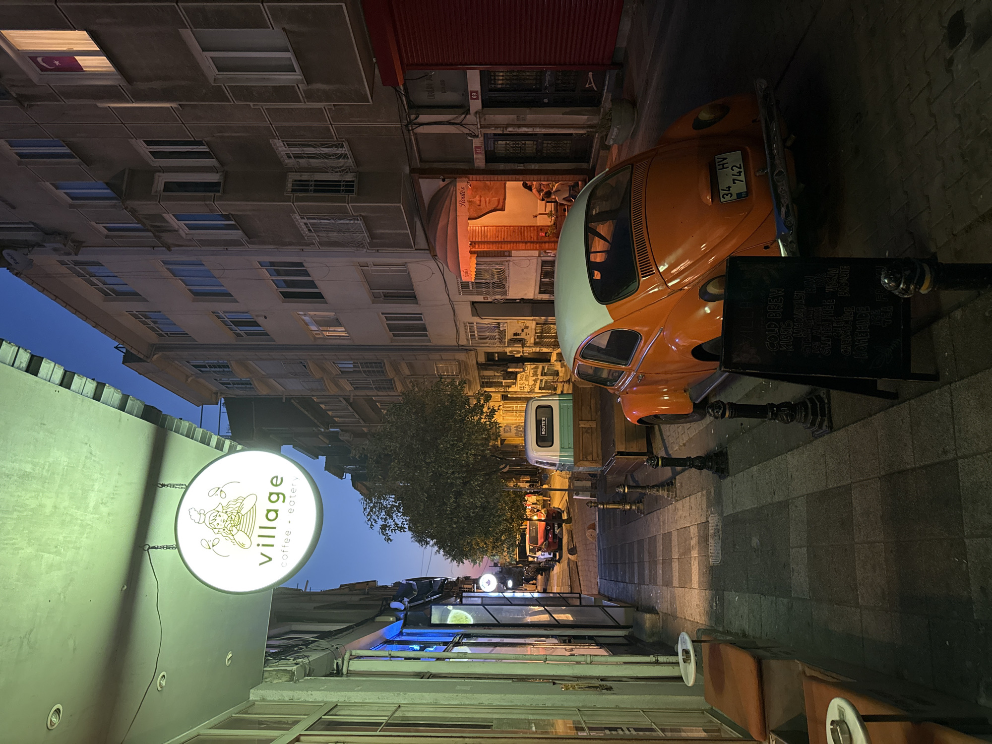 The image depicts a narrow street scene in Kadıköy, Turkey, at dusk. On the left, a cafe with a lit sign reading "village coffee + eatery" is visible, with outdoor seating provided by padded benches and small tables. The street is paved with cobblestones and is lined with buildings. An orange Volkswagen Beetle is parked on the right side of the street, with its license plate reading "34 HV 742". A chalkboard menu stands in front of the car. Further down the street, a light blue and white vintage truck is parked, and several other cars and people are visible. The sky is a deep blue, indicating the transition from day to night, and artificial lighting from the cafe and street lamps illuminates the scene. The buildings flanking the street are multi-story and appear to be residential or commercial. A Turkish flag is visible in a window of one of the buildings on the right.