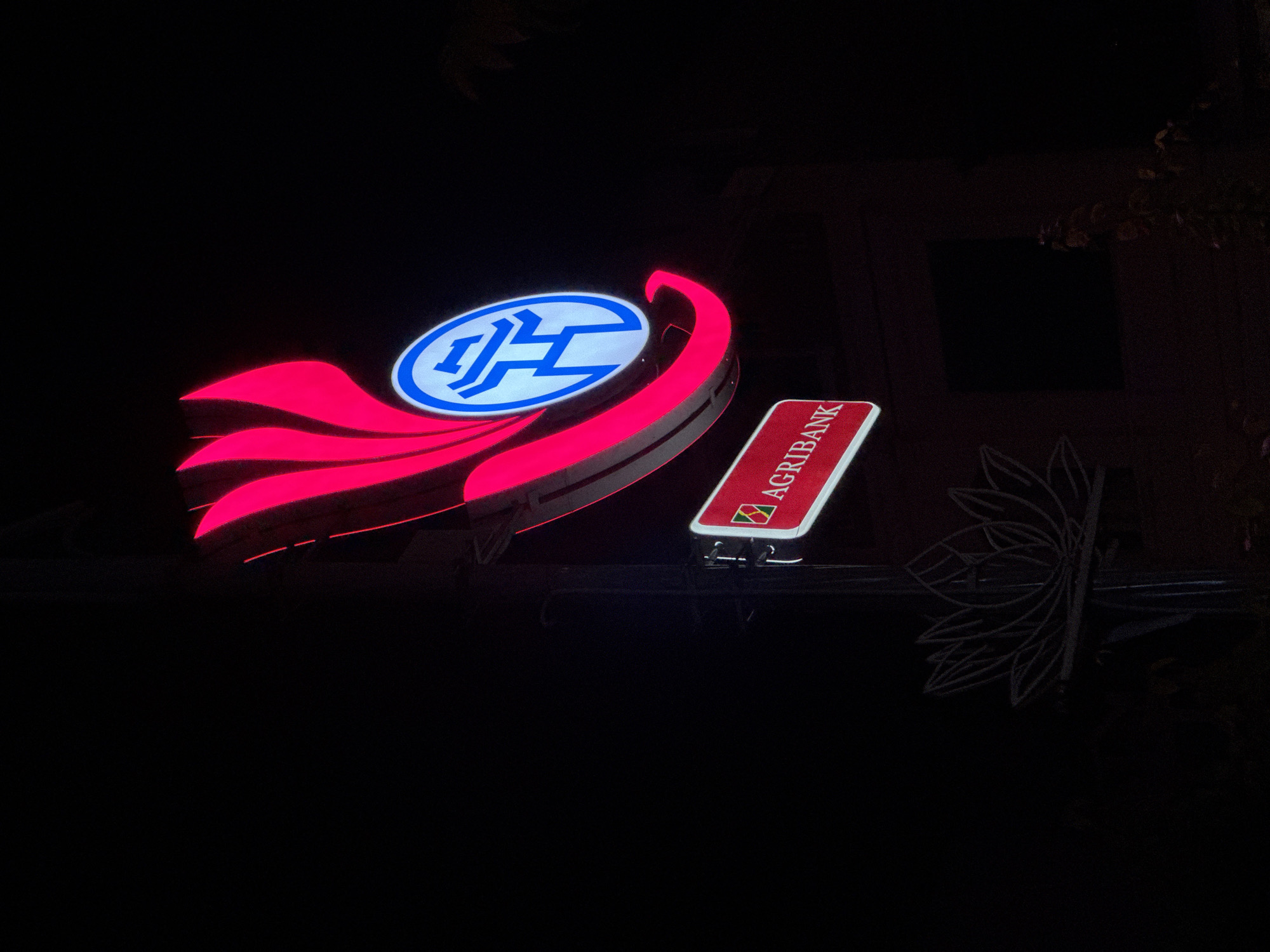 The image shows a neon sign for Agribank, a bank in Vietnam. The sign is illuminated in red and blue and is mounted on a pole. Below the Agribank sign, there is a decorative metal lotus flower. The sign is lit at night, suggesting it is in a city. The background is dark, with some hints of buildings and trees. The overall impression is that of a well-lit advertisement in a nighttime urban setting.