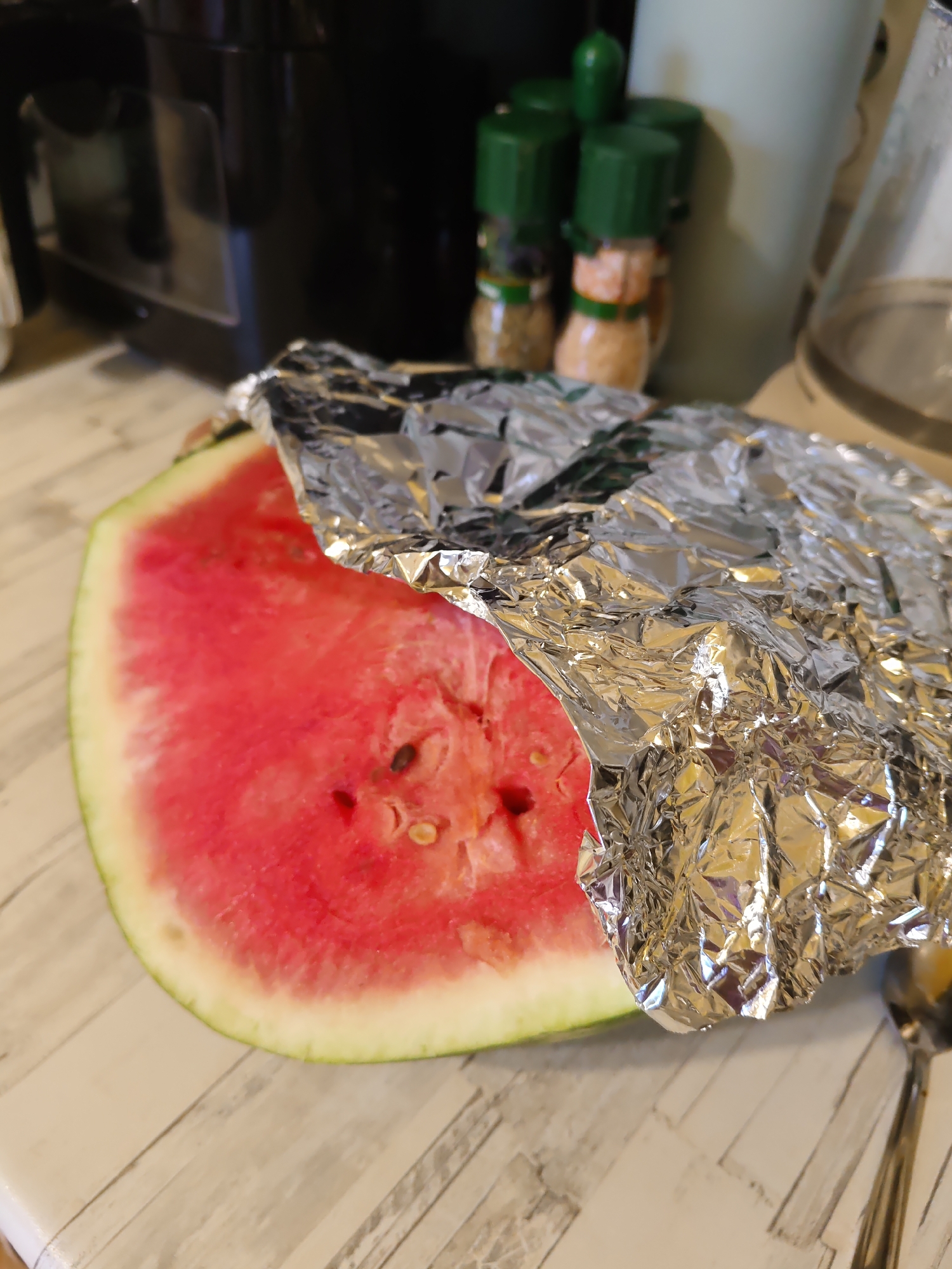A wedge of watermelon, partially covered in crumpled aluminum foil, rests on a light-colored wooden surface. The watermelon is bright red with visible seeds and a green rind. To the right of the watermelon, a metal spoon is partially visible. In the background, out of focus, are kitchen appliances, including a black appliance, spice shakers with green lids, and a tall, light-colored cylindrical object. This scene is located in a kitchen in Rostov-on-Don, Russia.