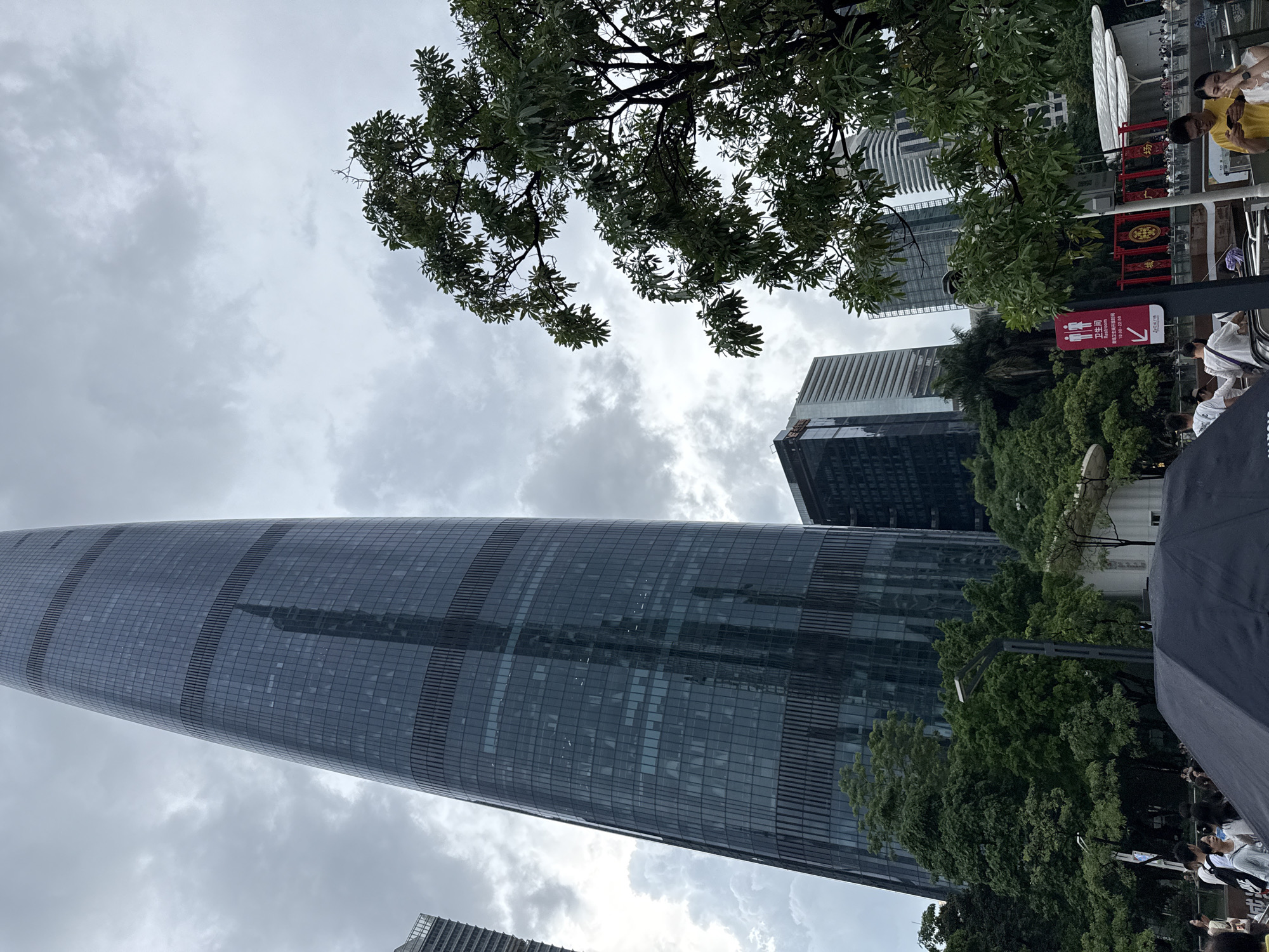 A wide shot captured on an overcast day shows a modern cityscape in Tianhe District, China. Dominating the left side of the frame is a very tall, sleek skyscraper with a dark, glass facade. Its surface reflects the cloudy sky and the surrounding buildings. To the right, a lush green tree with broad leaves partially obstructs the view of the sky and some of the buildings. In the mid-ground, several other contemporary buildings of varying heights are visible, including one with a distinct patterned facade. At the bottom of the image, a crowd of people are milling about, some are looking at their phones, while others are walking. In the foreground, a large black umbrella is open, obscuring the bottom portion of the scene. A red sign with pictograms and Chinese characters is visible on the right, near the crowd. The overall atmosphere is that of a busy urban environment on a cloudy day.