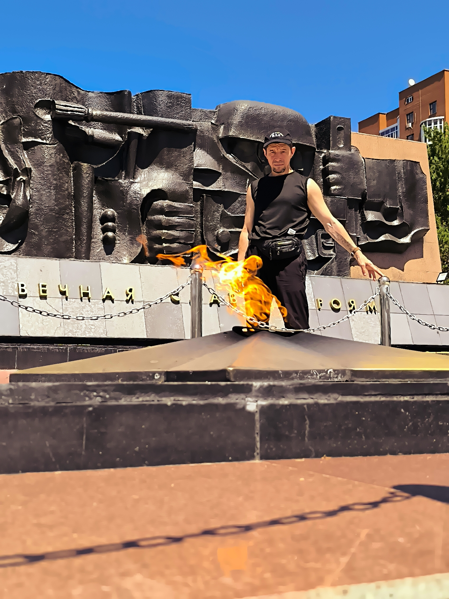 A man stands in front of an eternal flame monument in Qarağandı, Kazakhstan. The monument features a bronze bas-relief with chains and inscribed text. The man is wearing a black tank top, black pants, a black baseball cap, and a fanny pack. He is reaching out towards the monument with his right hand. The flame is lit and is a prominent feature in the foreground. The background shows a bright blue sky and buildings.