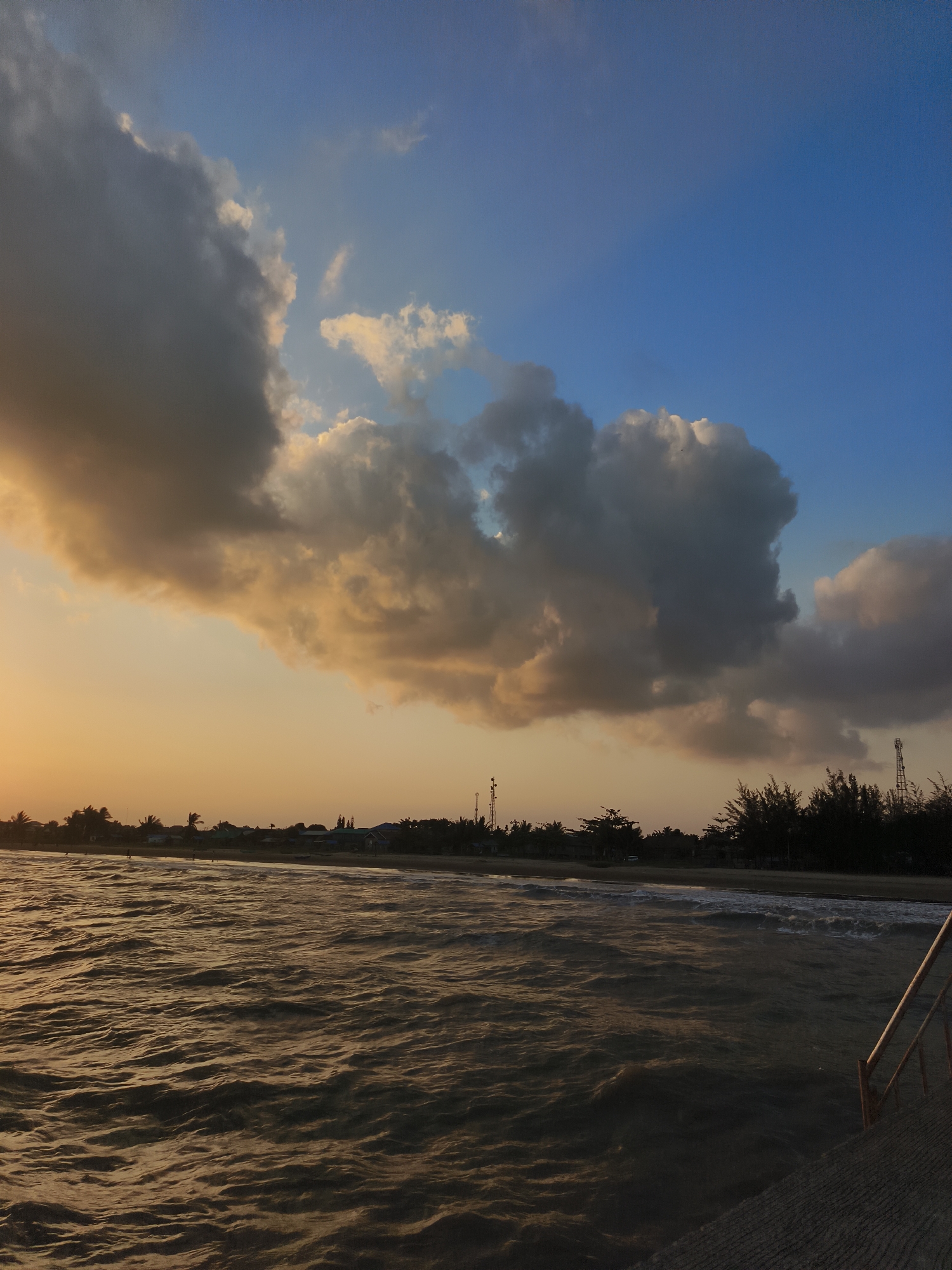 The image depicts a seascape at dusk in Kupang Berkah Jaya, Indonesia. The sky is a gradient of warm orange and yellow near the horizon, transitioning to a deep blue at the top, with scattered dark and light clouds. The sea is choppy, with visible waves reflecting the warm hues of the sky. In the distance, a shoreline is visible with a line of trees and a few buildings, including what appears to be a tall communication tower. The foreground shows a concrete structure with a metal railing, suggesting a pier or walkway. There are no identifiable people or specific actions captured in the frame.