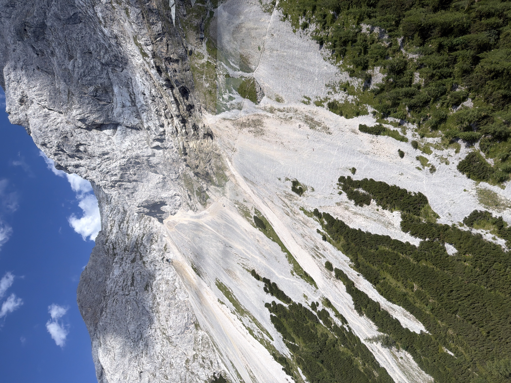 This media file captures an aerial view of a dramatic mountainous landscape in Grainau, Germany. The scene is dominated by a towering, sheer rock face on the left, its textured surface displaying layers of grey rock and shadowed crevices. In contrast to the starkness of the cliffs, the right side of the image reveals a steep slope covered in dense, dark green vegetation, appearing as thick bushes or low-lying trees.
Between the cliff face and the vegetated slope, a vast expanse of scree or gravel stretches across the mountain, marked by lighter grey tones and occasional patches of sparse green growth. A faint, winding path can be discerned on this scree slope, suggesting a hiking trail or access route. The sky is a vibrant blue, with a few scattered white clouds adding depth to the scene. The lighting suggests it is daytime, likely late morning or early afternoon, with the sun casting shadows that highlight the rugged terrain.
The overall impression is one of natural grandeur and remoteness. The sheer scale of the rock face and the steepness of the vegetated slopes emphasize the wild and untamed nature of the alpine environment. There are no visible people or man-made objects, besides the implied trail. The image conveys a sense of awe and the power of nature.