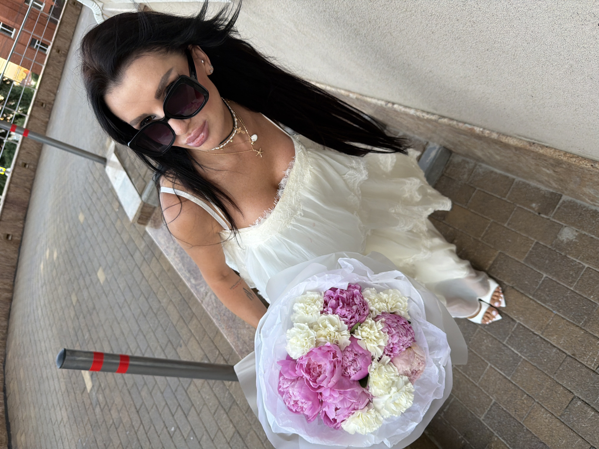 A woman with long, dark hair and sunglasses stands on a patterned brick walkway with a light-colored concrete wall to her right. She is wearing a white, lace-trimmed dress and white heeled sandals. She is holding a bouquet of pink and white peonies and carnations wrapped in white paper. A metal bollard with red stripes is visible to her left. In the background, a building with windows and a railing can be seen. The scene appears to be in an outdoor urban setting. This is the city of Novaya Usman, Russia.