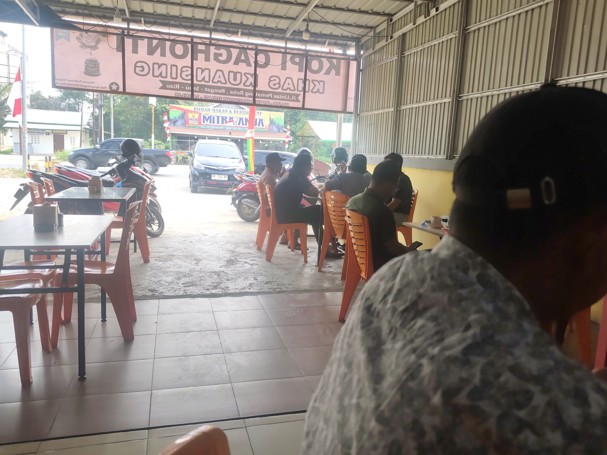 The image shows the exterior of a roadside cafe in Pematang Reba, Indonesia, with a corrugated metal roof supported by metal beams. Several motorcycles are parked outside, and the cafe is furnished with orange plastic chairs and metal tables. In the foreground, a person is seated, facing away from the camera. In the background, a black car and a pickup truck are visible on the road. A banner displaying text in Indonesian hangs above the entrance. A red and white Indonesian flag is visible on the left side of the frame. The cafe appears to be open for business.