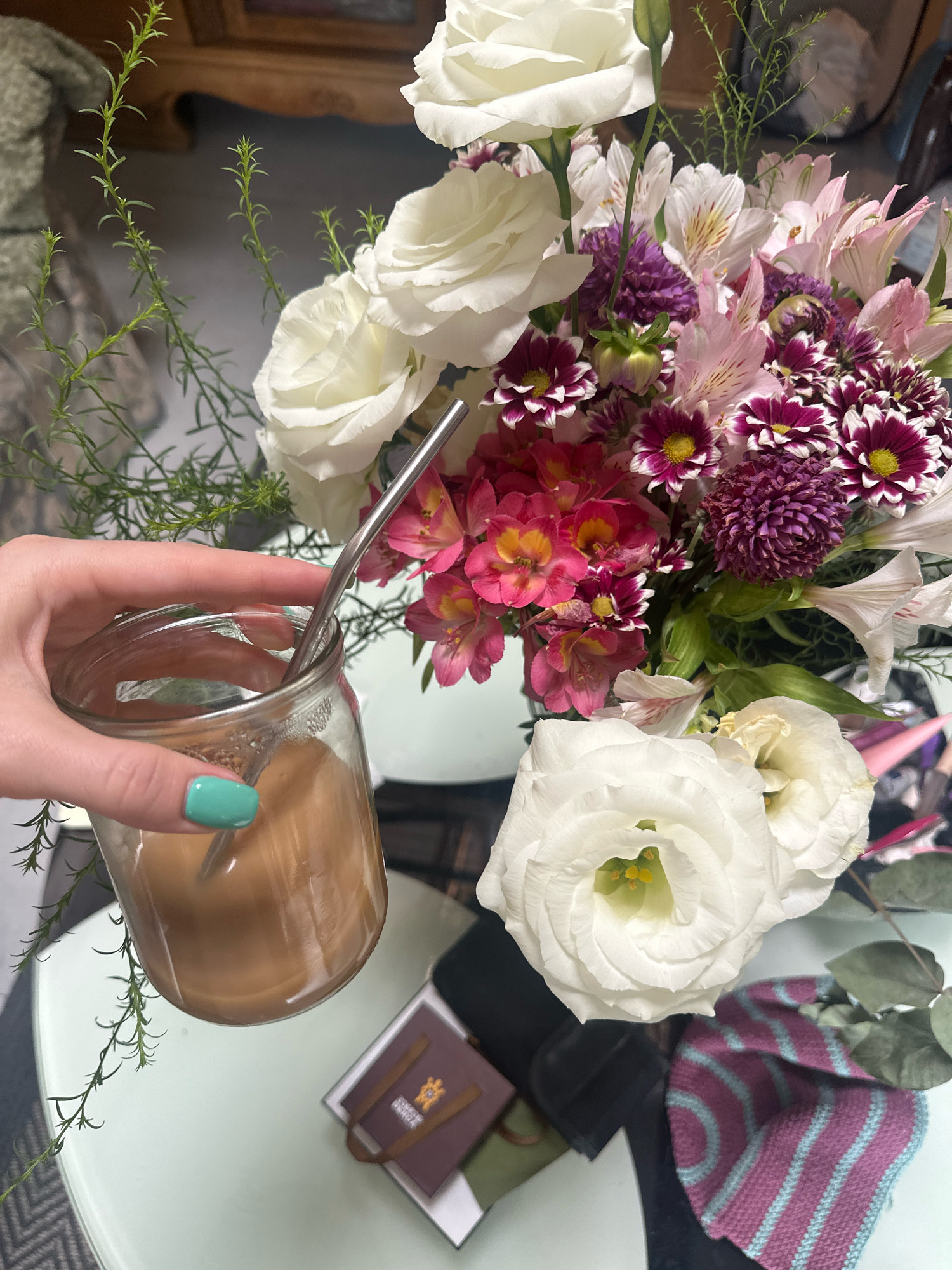 A hand with teal nail polish holds a clear glass jar filled with a light brown liquid and a metal straw. The jar is positioned in front of a bouquet of mixed flowers, predominantly white, pink, and purple, with some greenery. The flowers are arranged in a vase, and some are partially visible in the foreground and background. Several small brown gift bags are stacked on a table beneath the flowers. The table surface is light green and appears to be a round coffee table. A portion of a patterned rug is visible in the lower left corner of the image. The background shows a wooden piece of furniture with intricate carvings, suggesting an indoor setting. The lighting indicates it is daytime. The scene is likely a domestic setting or a cafe in Tbilisi, Georgia.