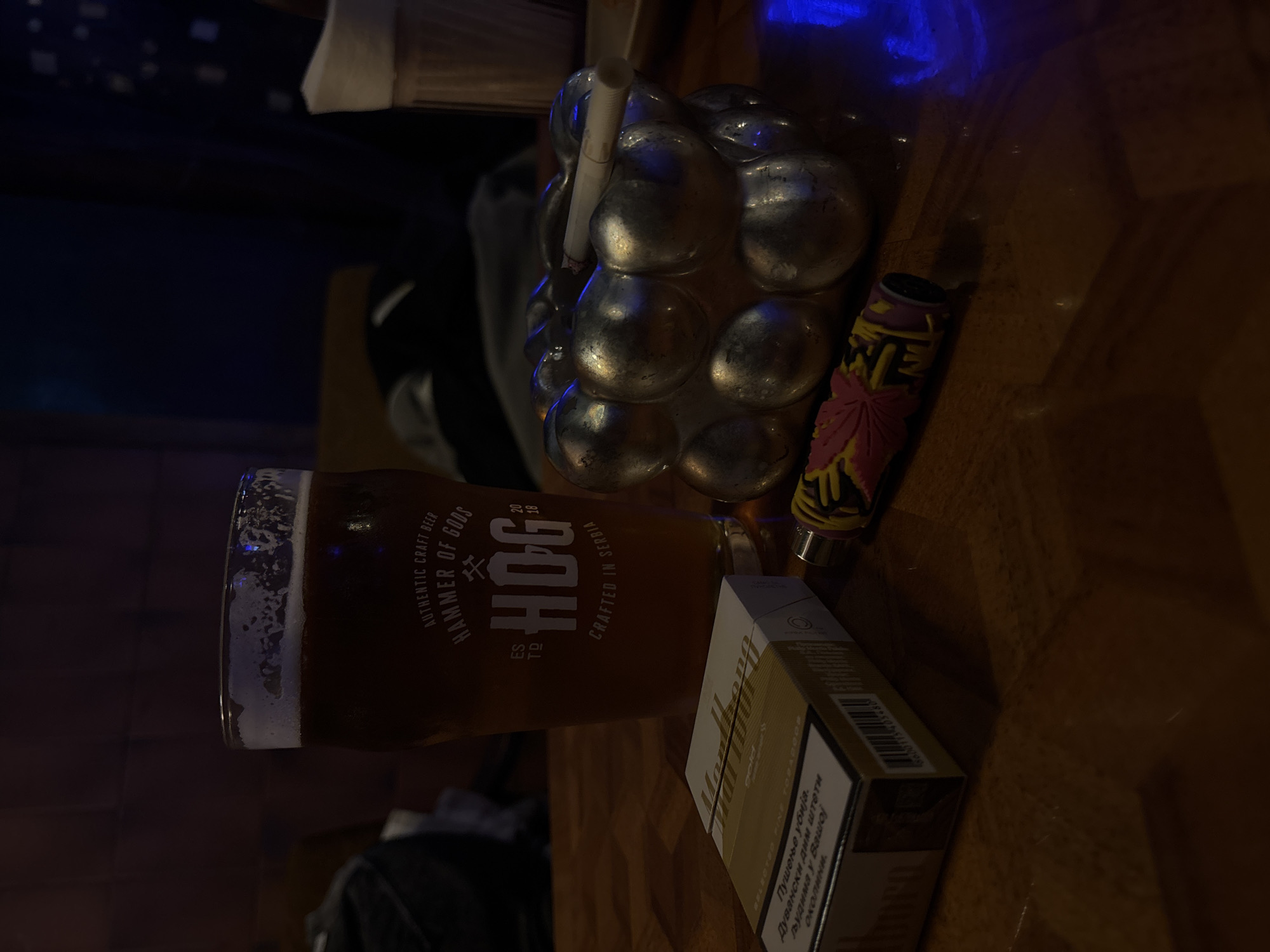The image shows a table setting with a glass of "Hammer of Gods" beer from Serbia, a pack of Marlboro Gold cigarettes with text in Serbian, a colorful lighter, and an ashtray holding a cigarette. The scene is likely in a bar or pub in Belgrade, Serbia, given the beer's origin and the Serbian text on the cigarette pack. The lighting is dim with some blue hues.